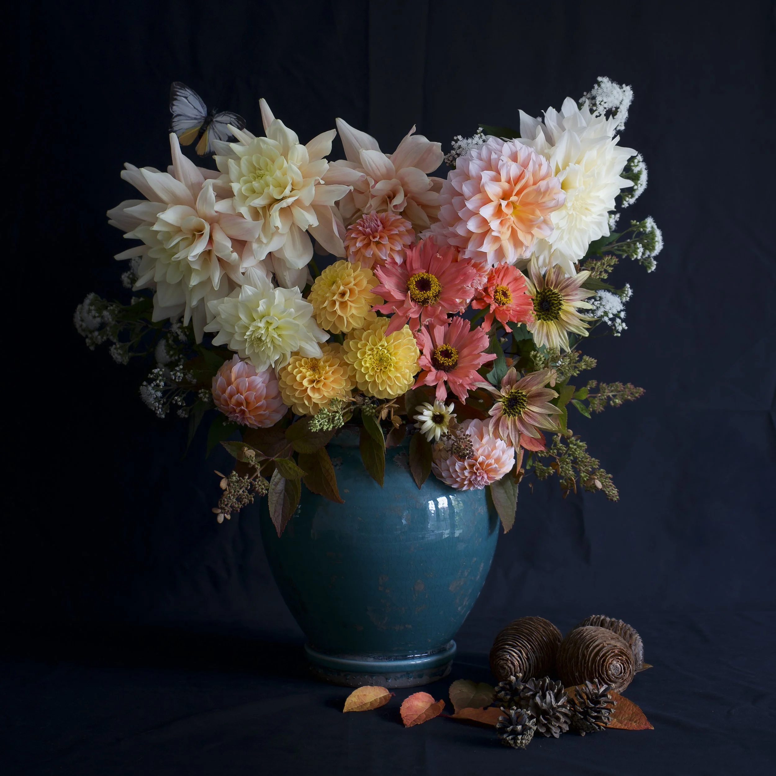A blue ceramic vase filled with a colorful arrangement of large dahlias, zinnias, and sunflowers, with a butterfly perched on a flower, set against a dark background, surrounded by pinecones and fall leaves.