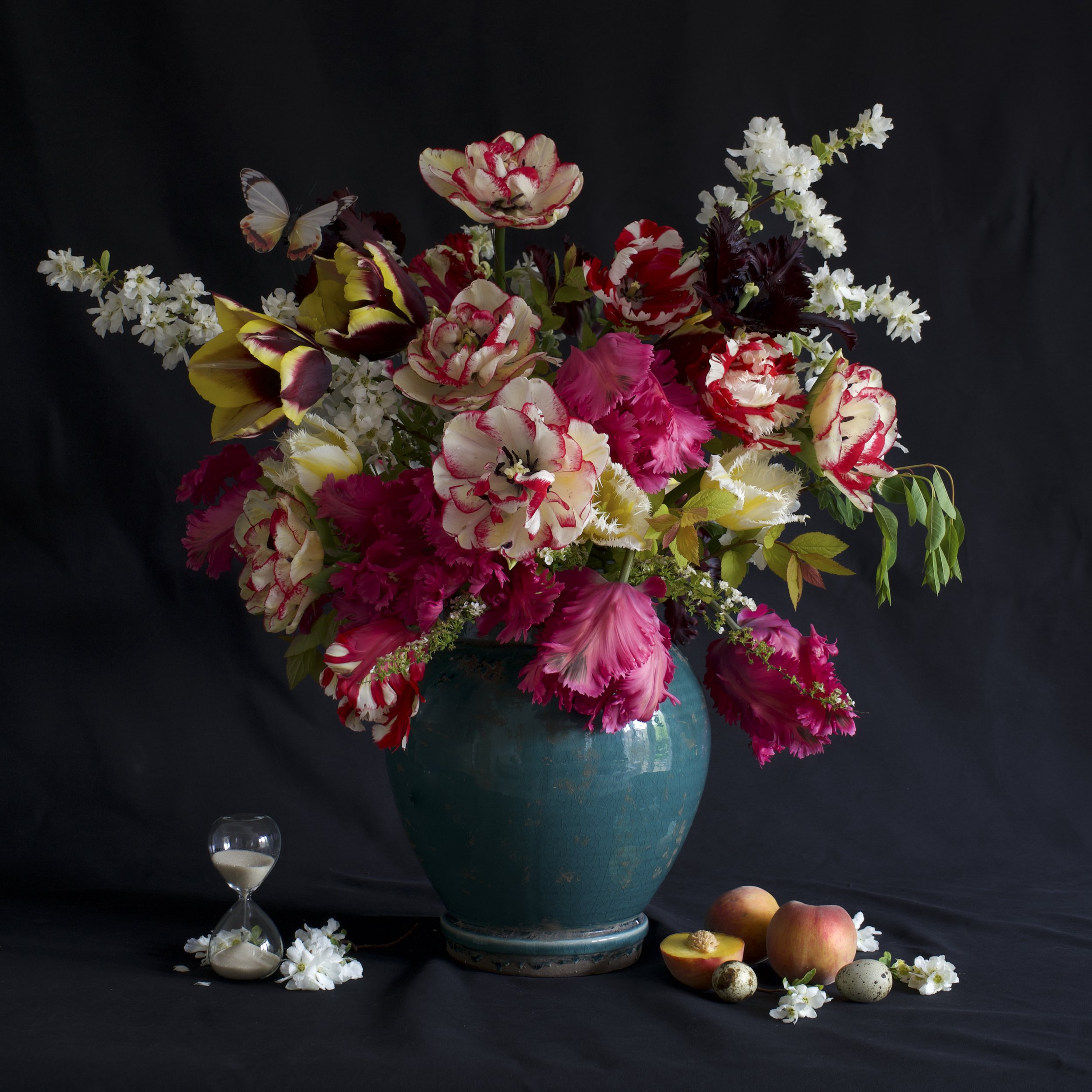 A large bouquet of colourful flowers in the style of still life in a blue ceramic vase including spring blossom and tulips, with fruit, bird eggs, and an hourglass on a black background.
