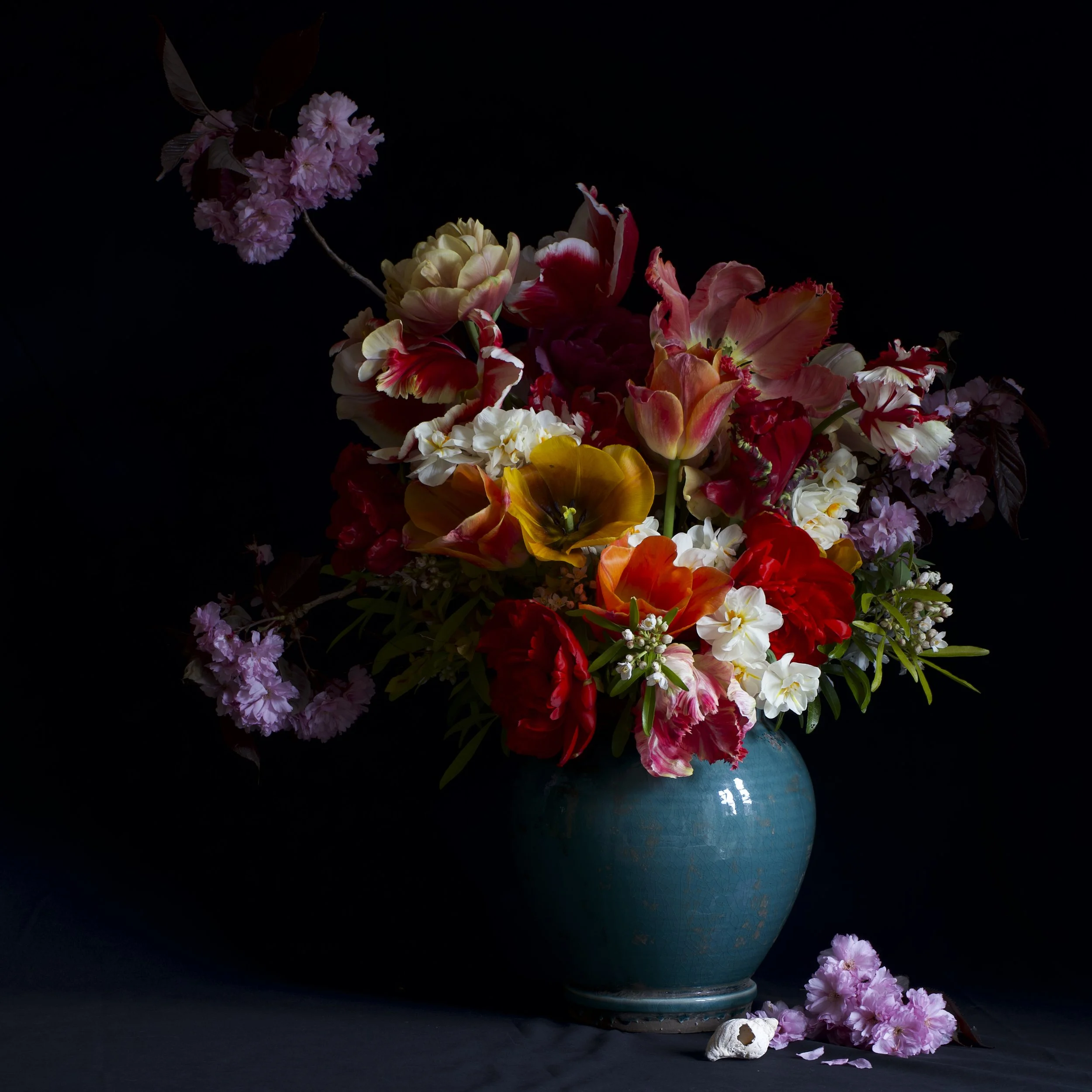 A vibrant bouquet of various colourful flowers in the style of still life including tulips, cherry blossom and narcissus in a blue ceramic vase, with some flowers and petals scattered at the base, against a dark background.