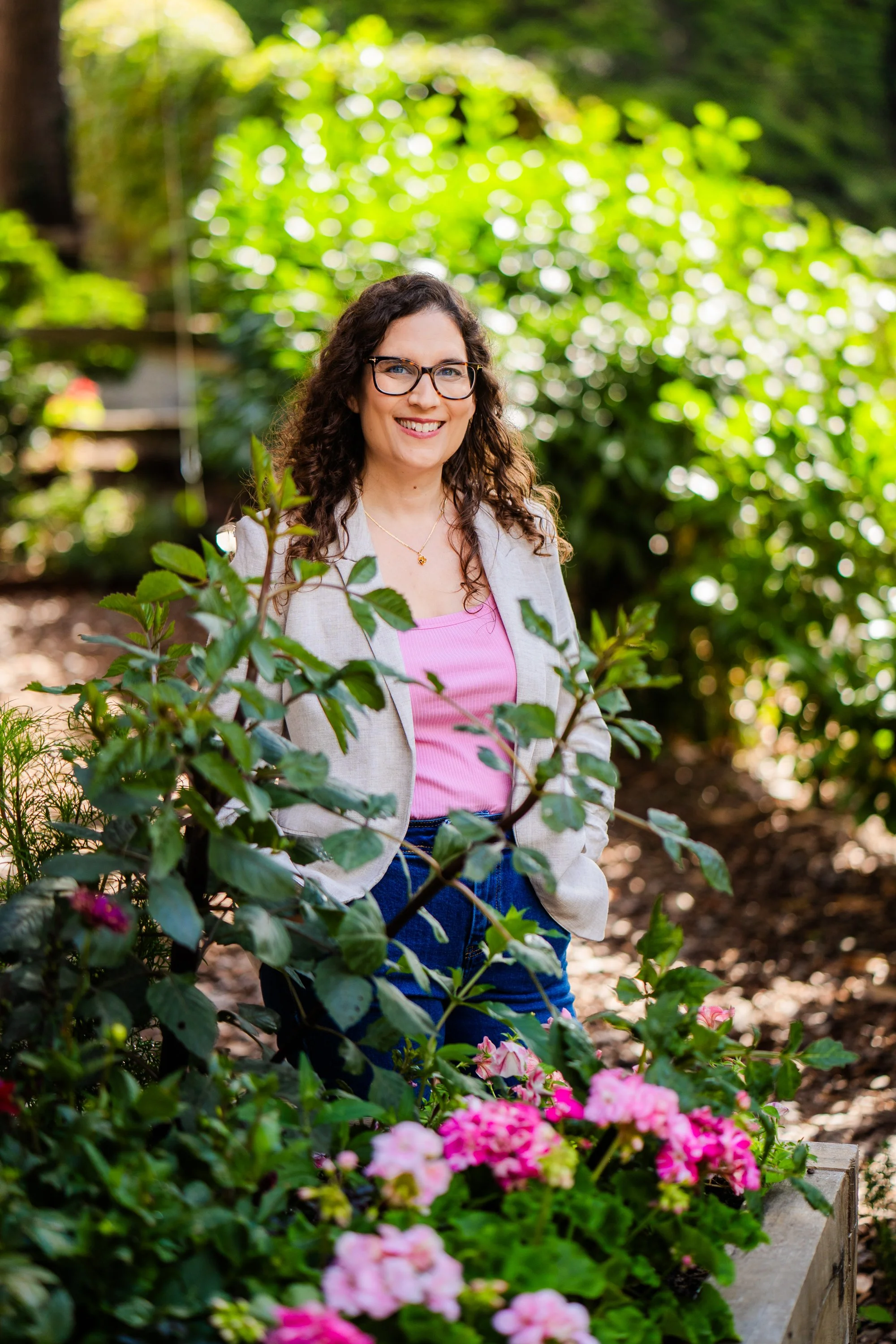 A woman with curly brown hair, glasses, and a bright smile standing outdoors among green plants and pink flowers.