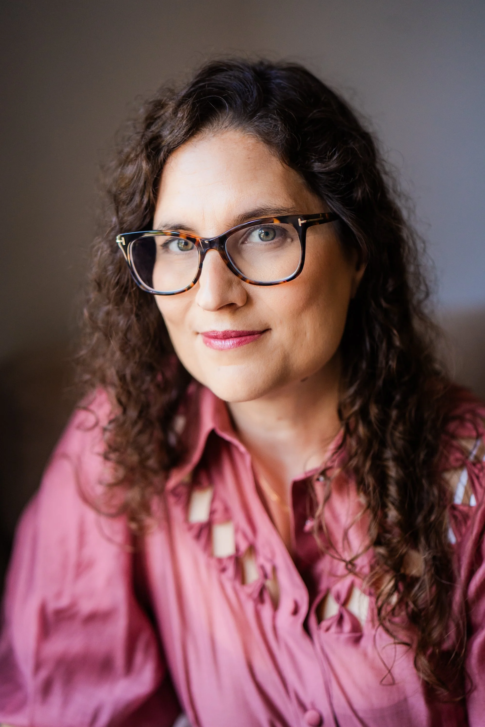 A woman wearing glasses with tortoiseshell frames and a pink blouse, looking at the camera with a slight smile, indoors with a neutral background.