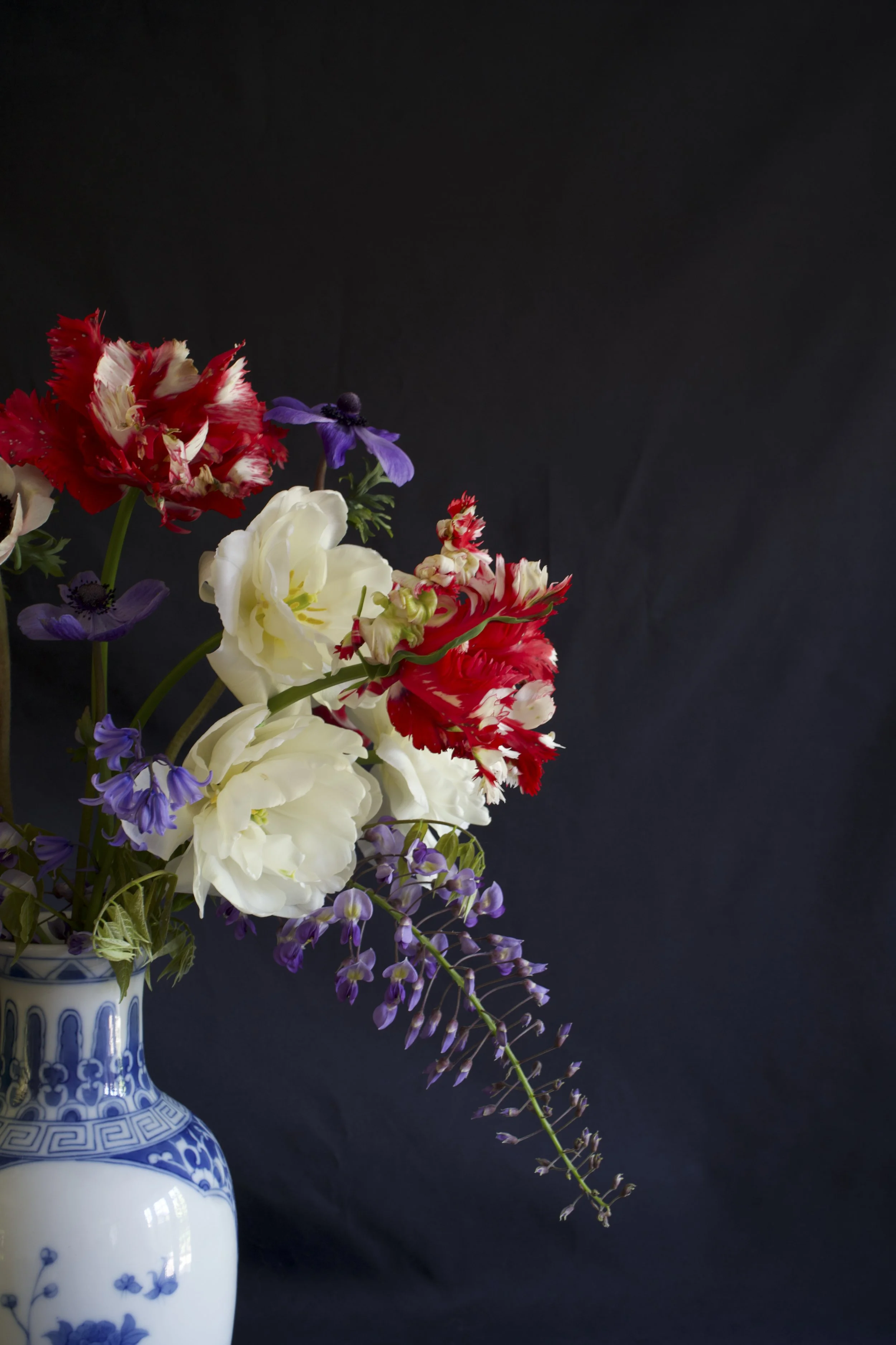 A bouquet of various colourful spring flowers including narcissus, tulips and wisteria in a blue and white patterned ceramic vase against a dark background.
