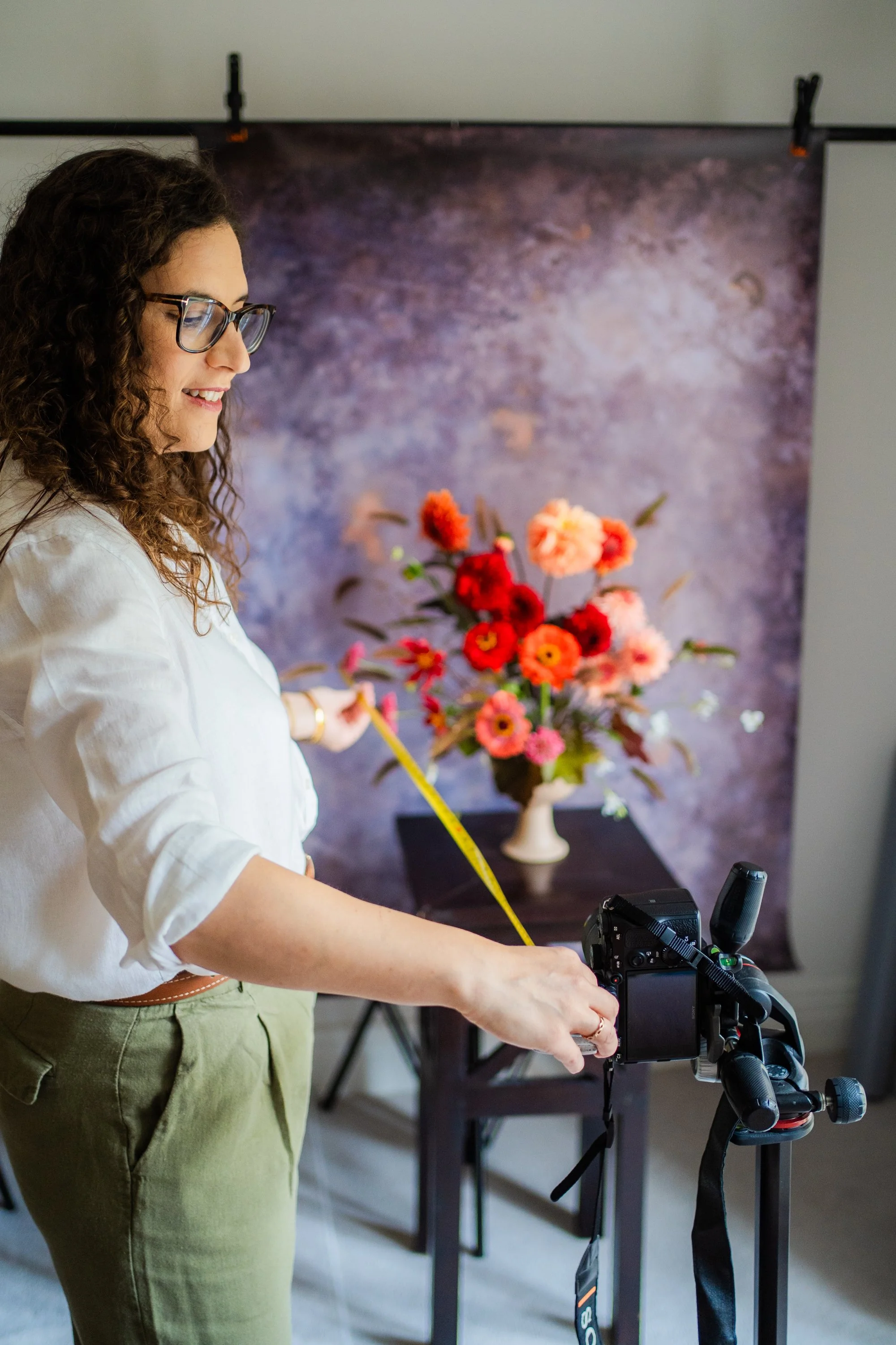 Woman taking a photo of a floral arrangement in a studio with a camera and measuring tape.