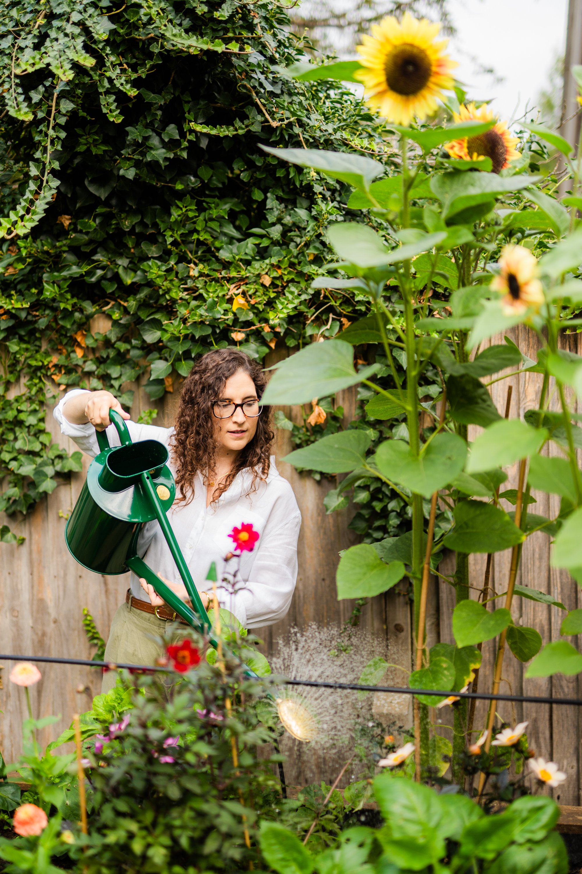A woman with curly hair and glasses watering plants with a green watering can in a cut flower raised bed garden with various flowers and a wooden fence, surrounded by green foliage.