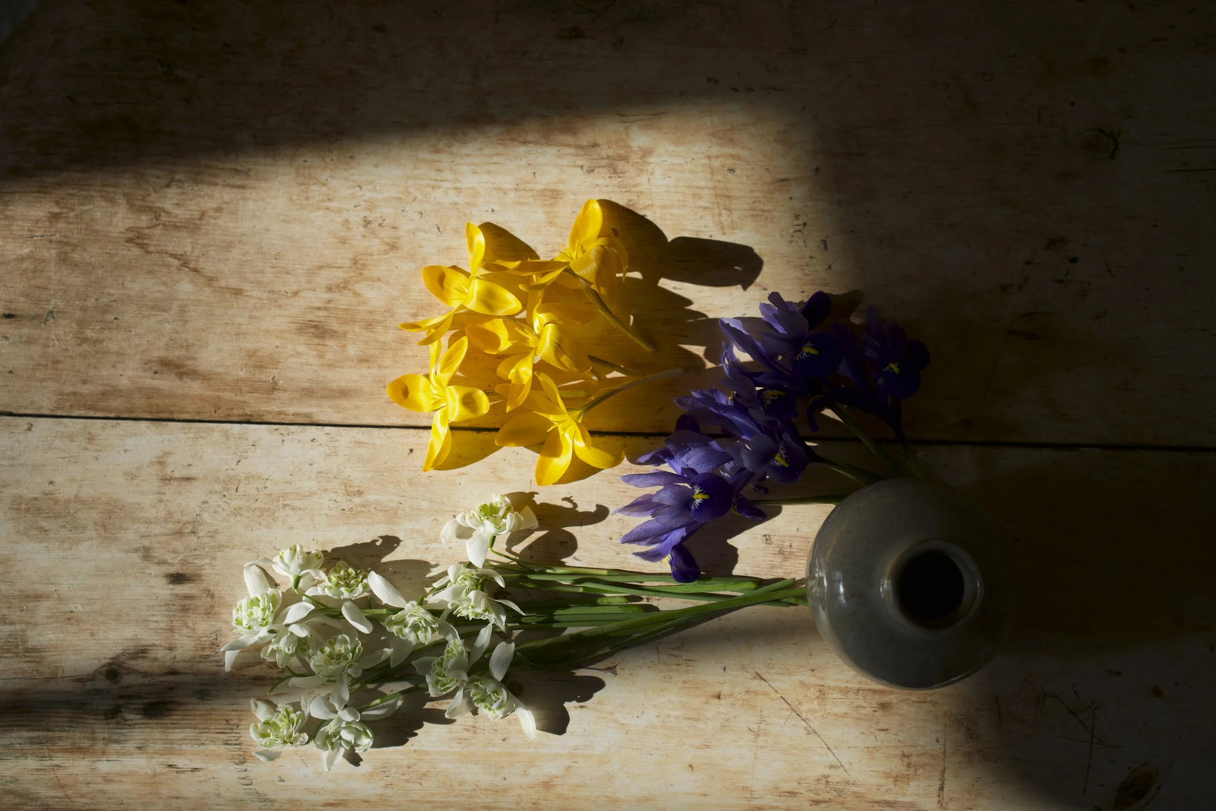 A bouquet of yellow crocus, purple muscari, and white snowdrop flowers  in a gray ceramic vase, placed on a wooden surface with sunlight casting shadows.