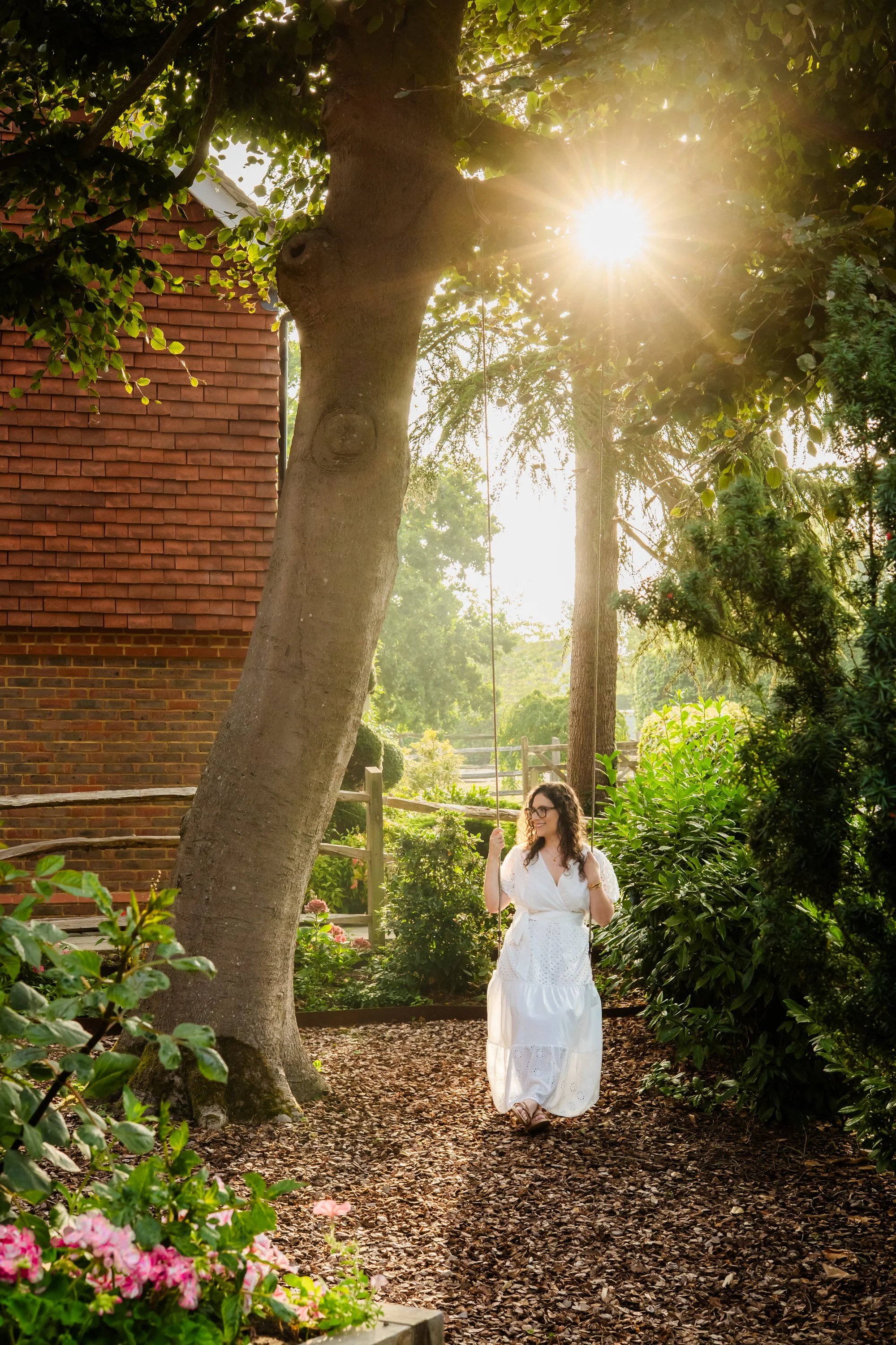 A woman in a white dress is swinging on a swing attached to a large tree in a lush garden with sunlight filtering through the leaves.