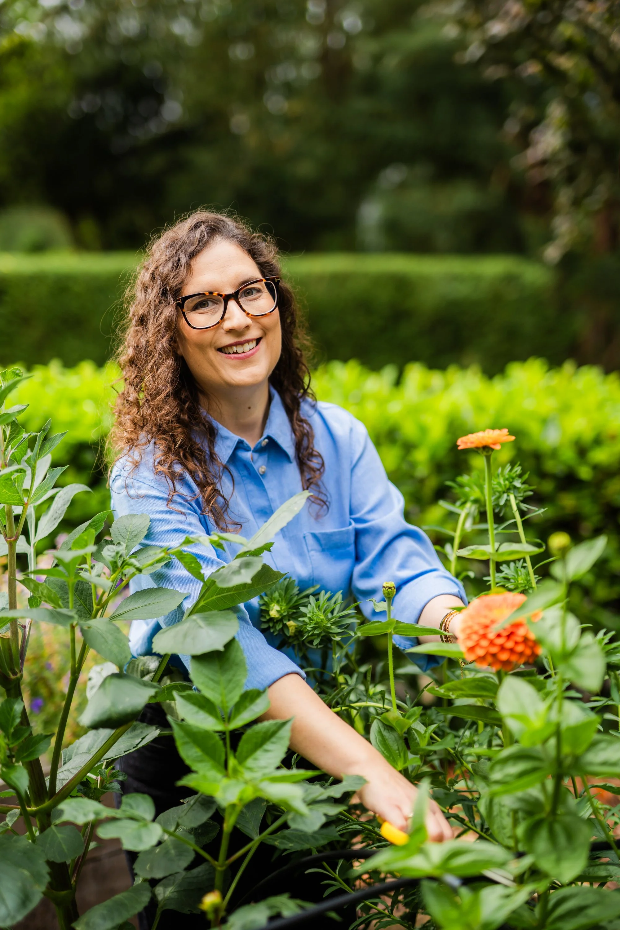 A woman with curly hair and glasses smiling while gardening among orange dahlias and green plants in a lush outdoor setting.