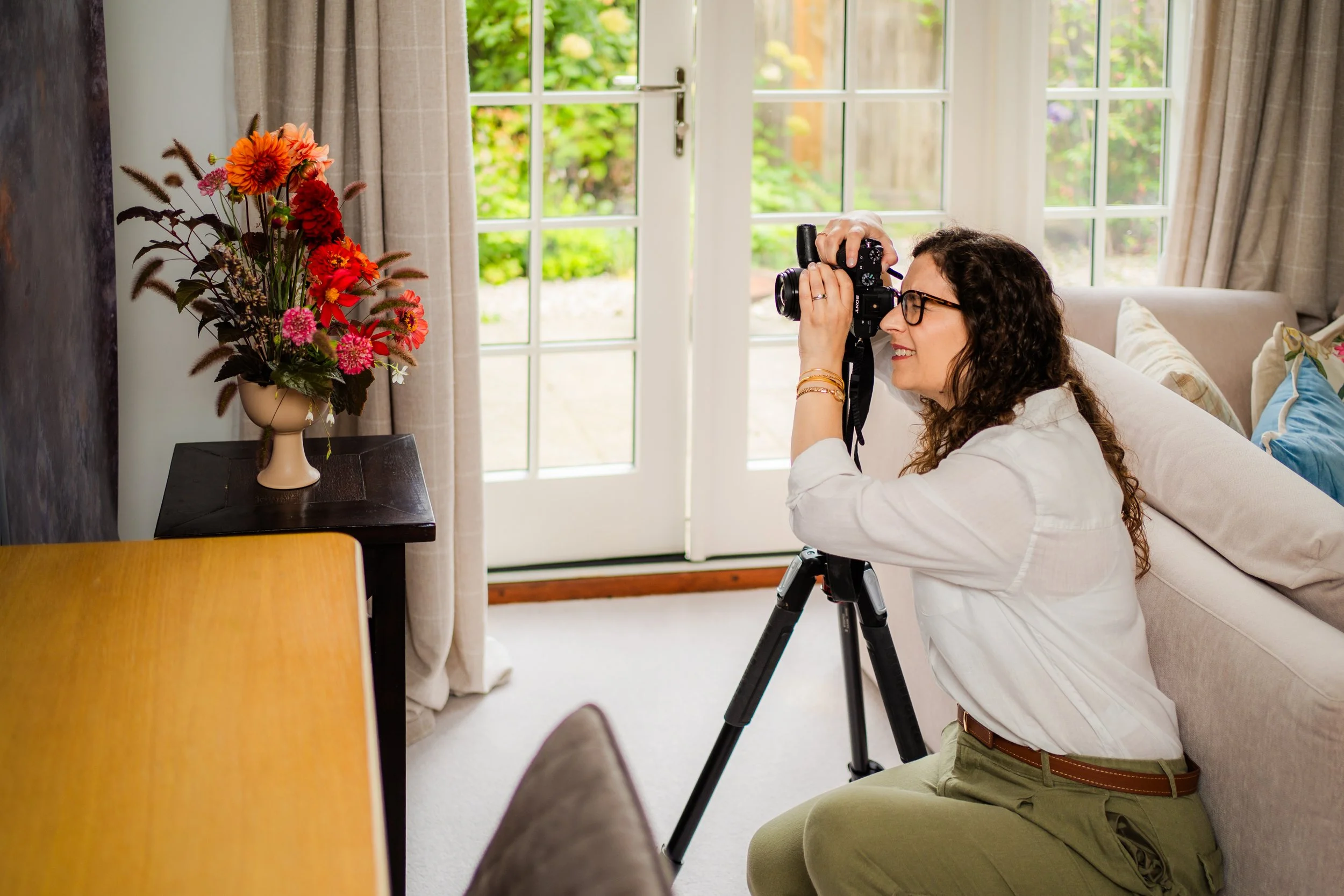 Woman with curly hair and glasses taking a photograph of flowers indoors with a camera mounted on a tripod, sitting on a beige sofa near a glass door with a garden outside, next to a small black table with a vase of colourful flowers.