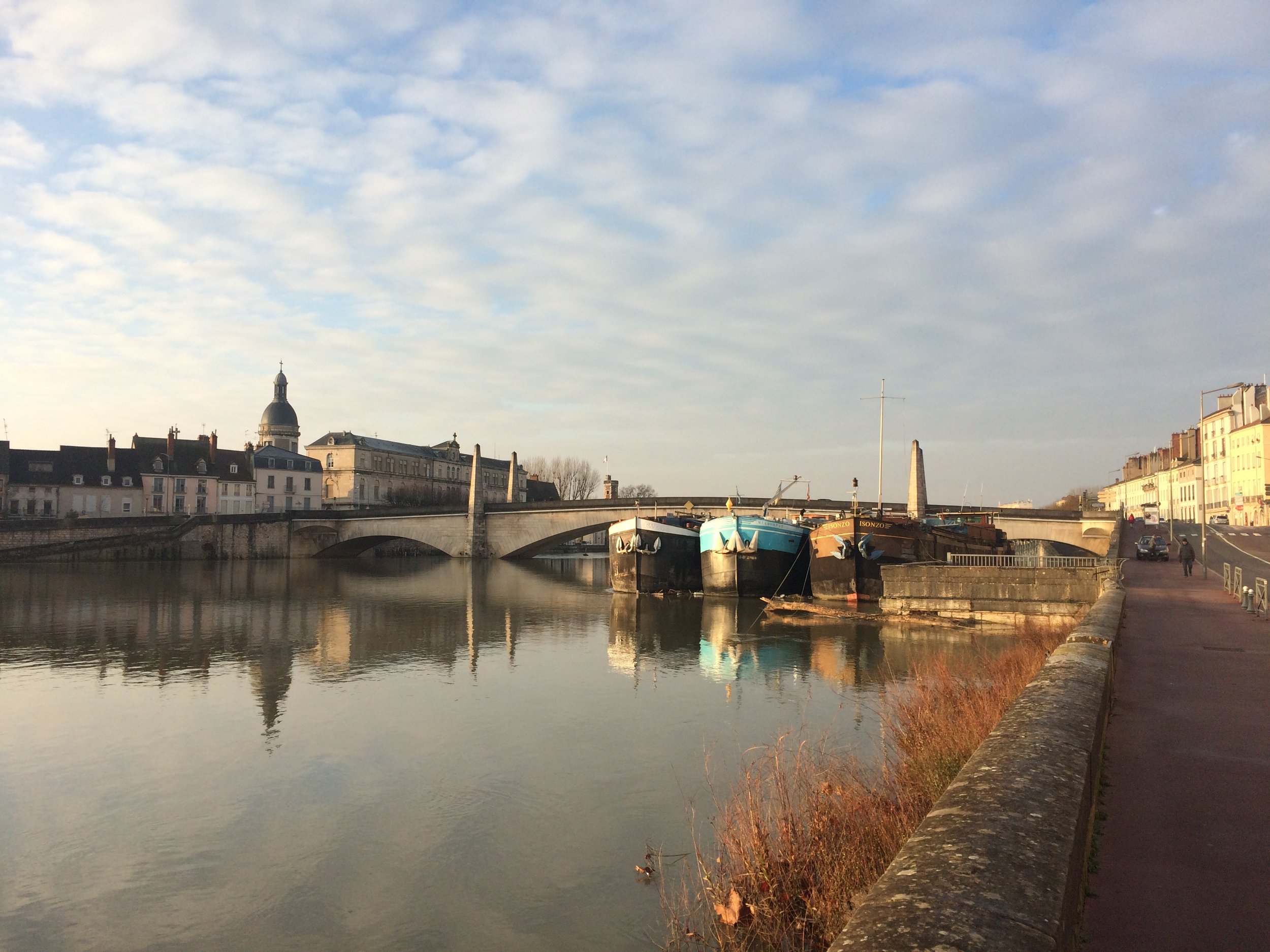 Chalon sur Saône, vue d'une rivière avec des bateaux amarrés, un pont, des bâtiments et un ciel partiellement nuageux à l'aube ou au crépuscule.
