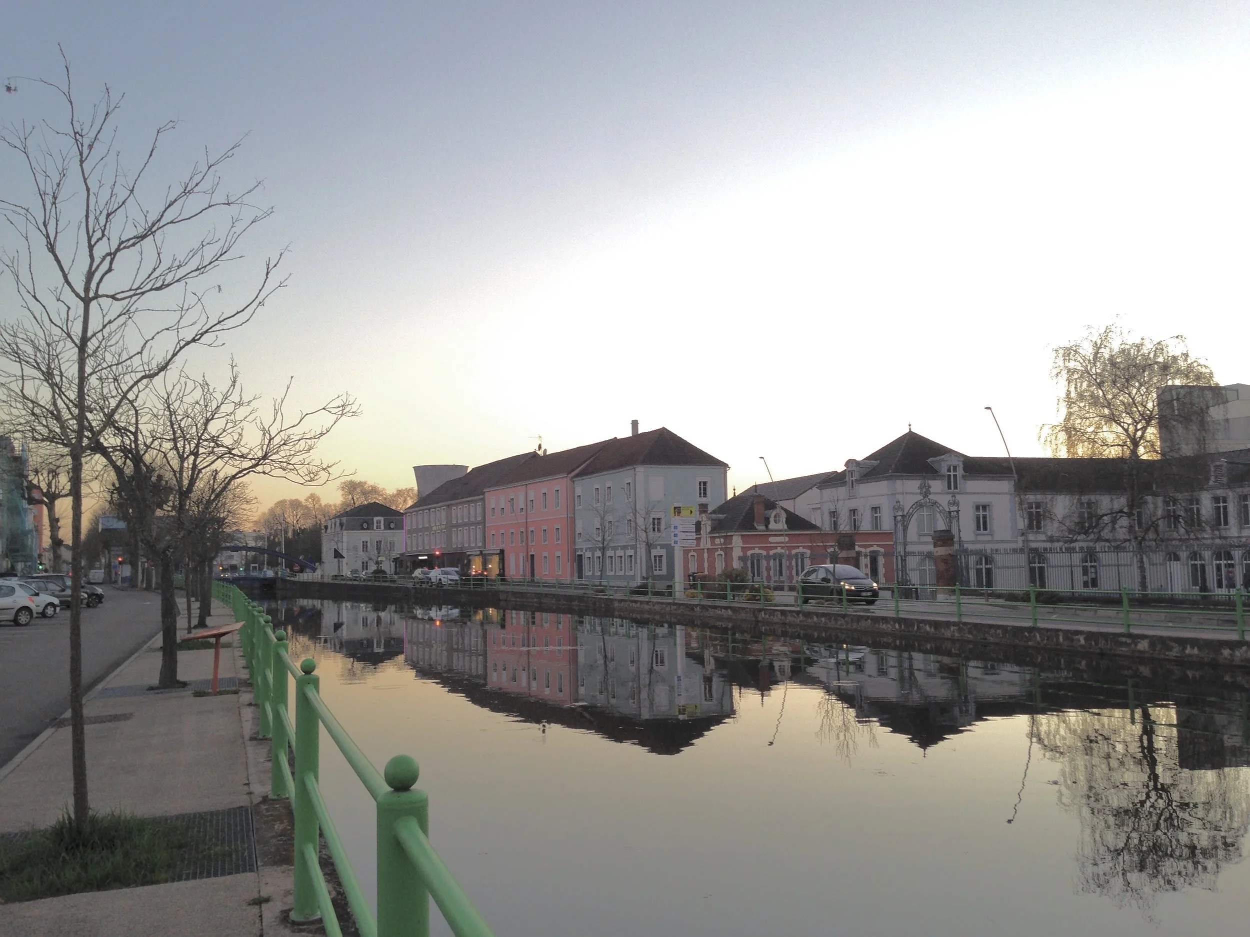 Vue d'une canal avec des bâtiments résidentiels colorés et des arbres nus, reflétés dans l'eau, au coucher du soleil.