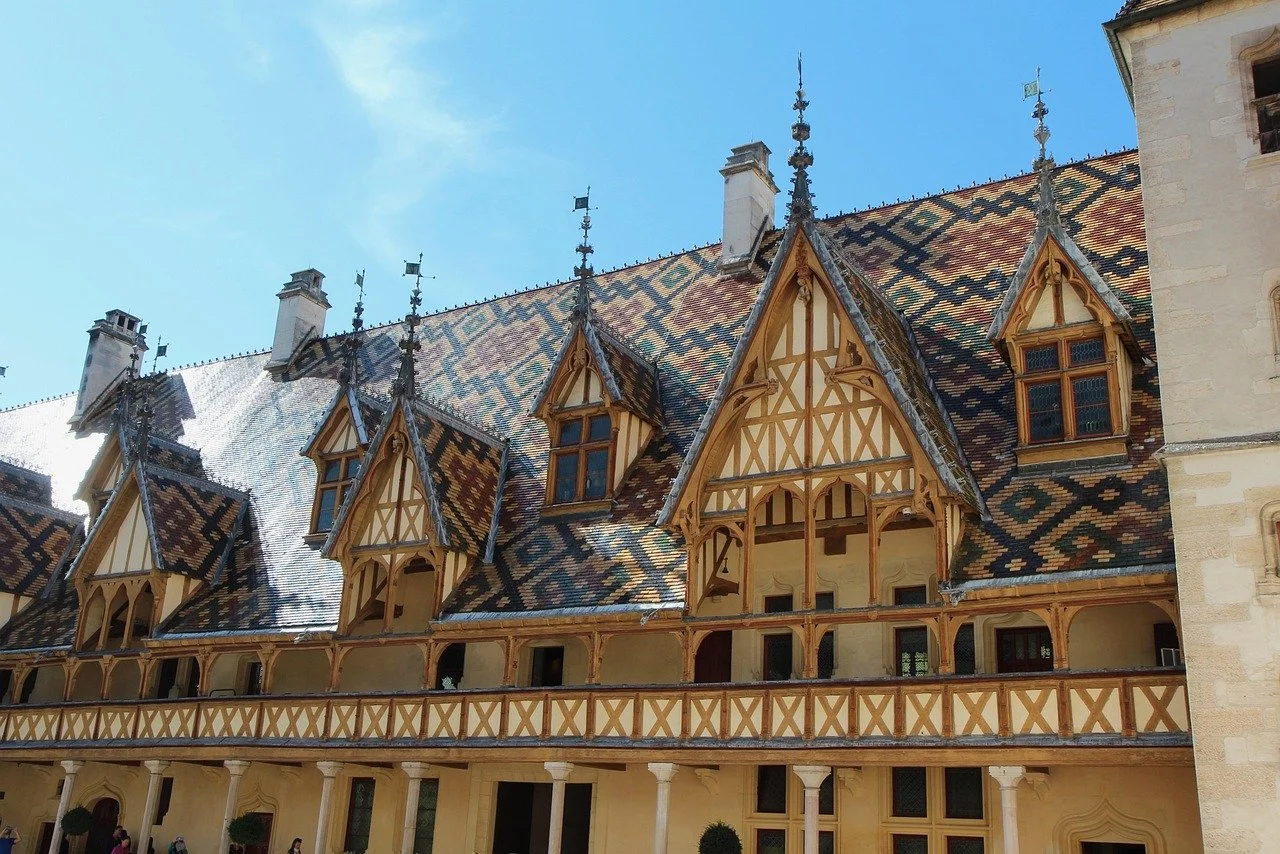 Vue d'un ancien bâtiment avec un toit en tuiles colorées et une architecture médiévale, comprenant des fenêtres à vitraux et une balustrade en bois.