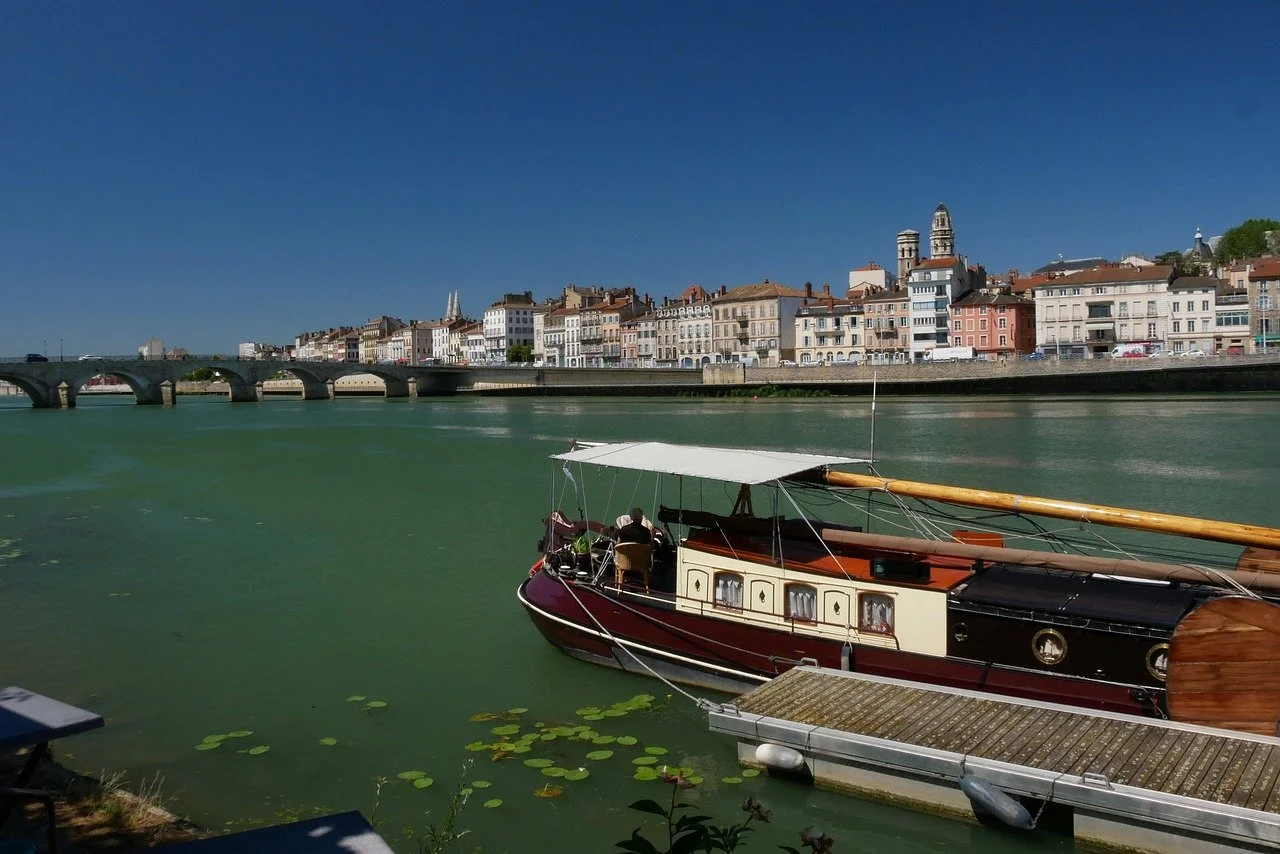 Un bateau à moteur amarré sur une rivière avec une ville historique en arrière-plan et un pont, sous un ciel bleu clair.
