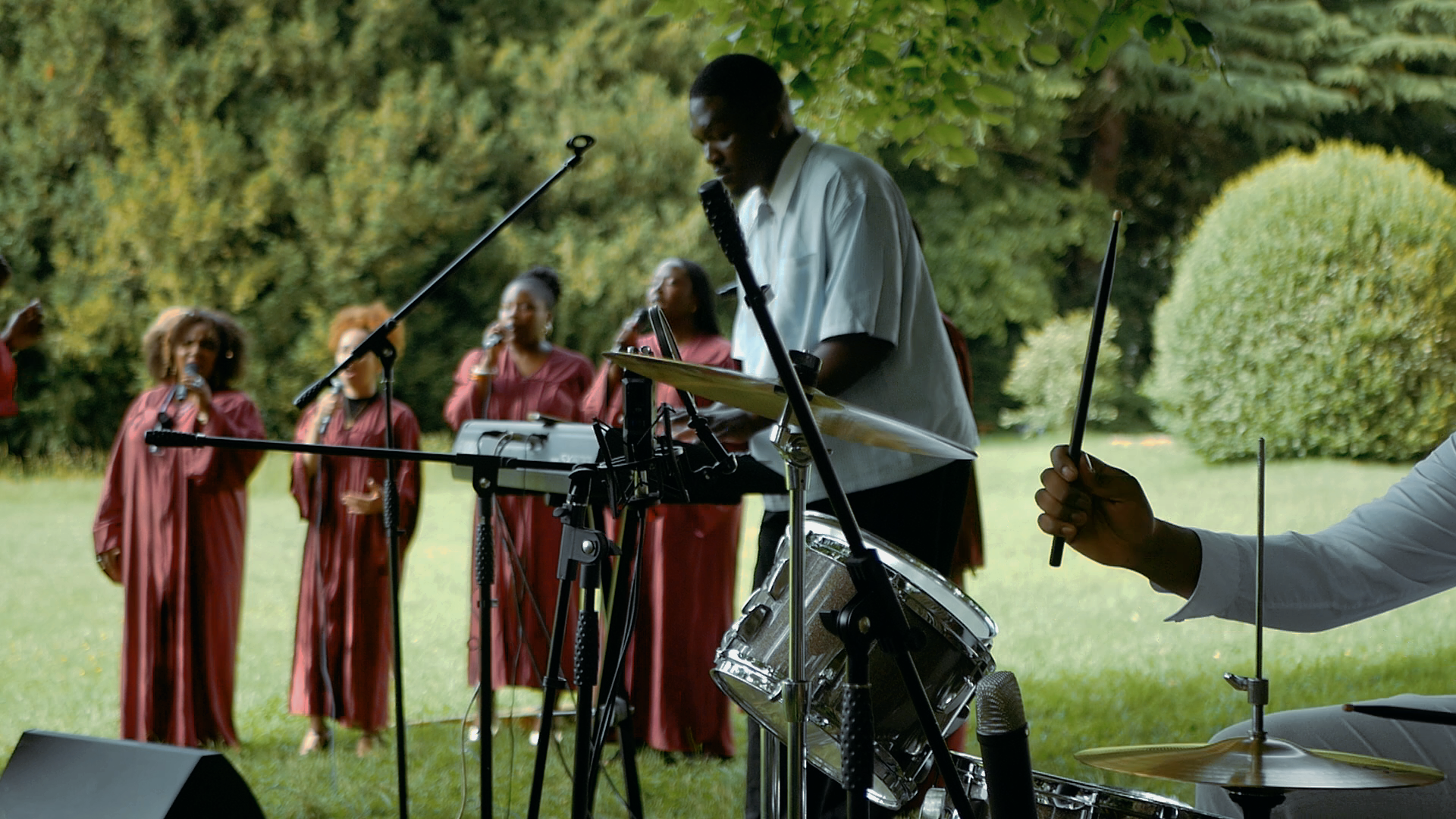 Un groupe de chanteurs vêtus de robes marron en train de chanter lors d'une performance en plein air, accompagnés de musiciens jouant de divers instruments. Le fond est constitué d'arbres et de végétation.