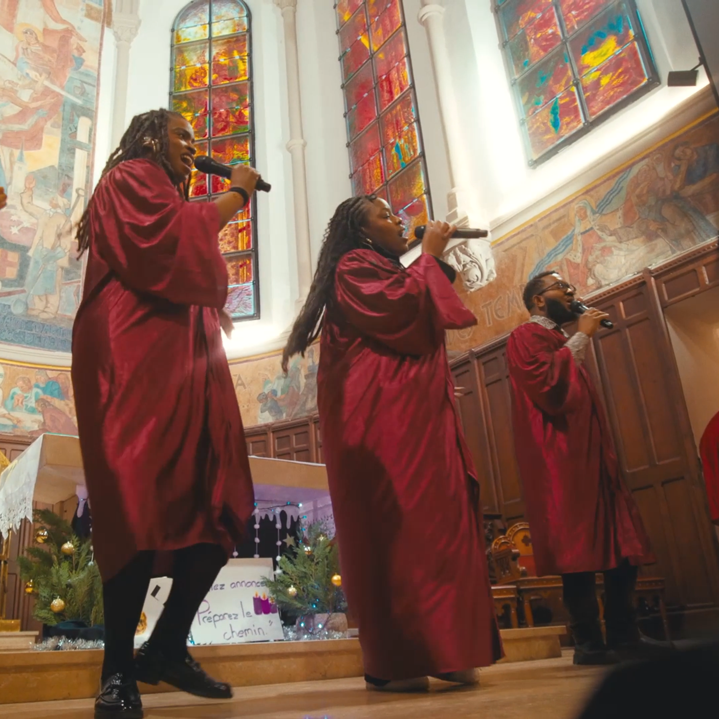 Trois personnes chantent avec des microphones dans une église, portant des robes rouges, avec des vitraux colorés en arrière-plan.