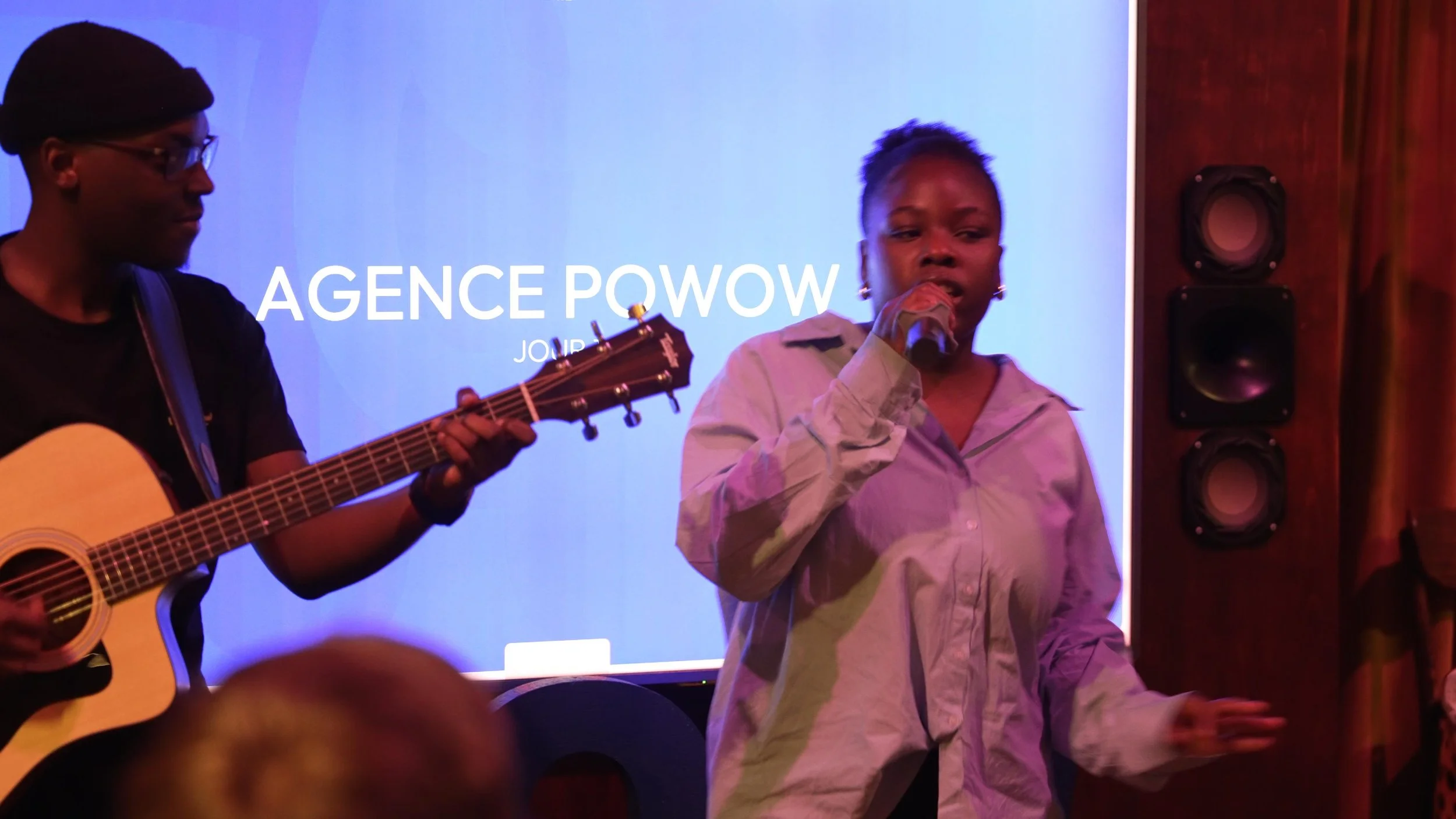 fête musical d'entreprise La femme à gauche joue de la guitare, la femme à droite chante dans un microphone. Le fond affiche le texte 'AGENCE POWOW'.