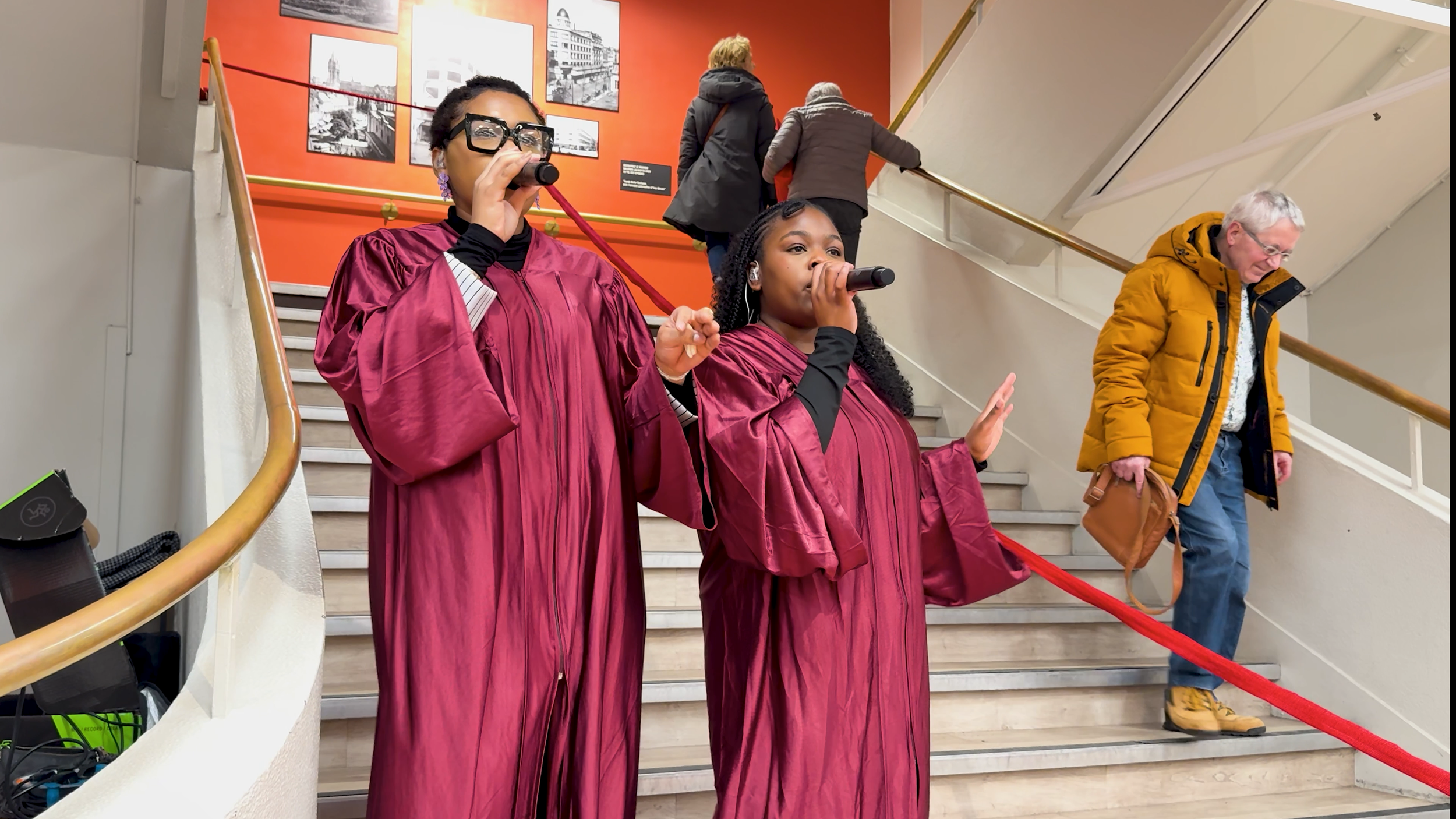 Deux femmes habillées en robes de cérémonie bordeaux toges du groupe Webelieve chantent avec un microphone dans une main, sur des escaliers, lors d'un événement aux Galeries Lafayette . D'autres personnes montent les escaliers en arrière-plan.