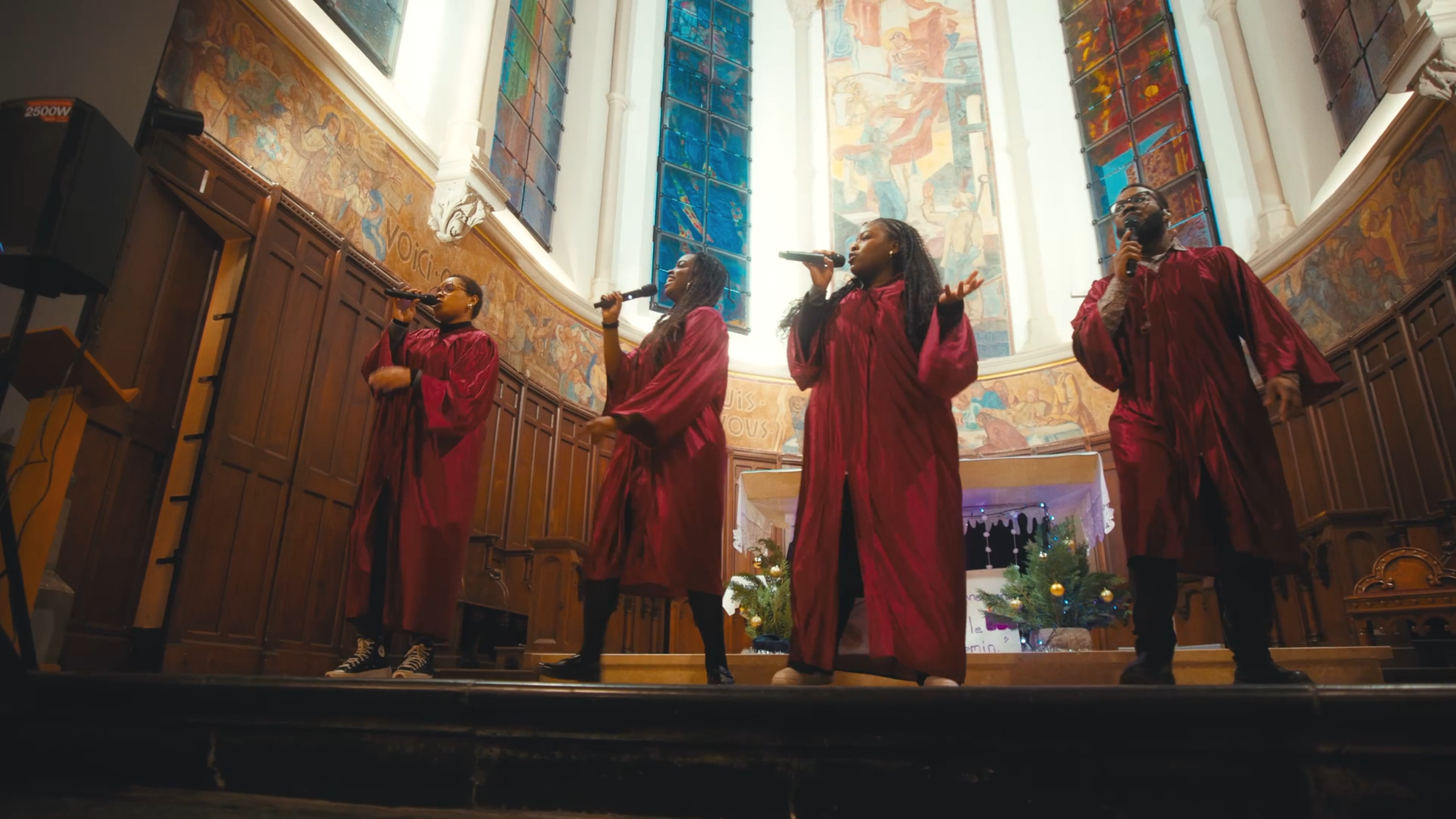 Quatre personnes chantent avec des microphones dans une église, toutes portent des robes de cérémonie rouges, avec des vitraux colorés en arrière-plan.