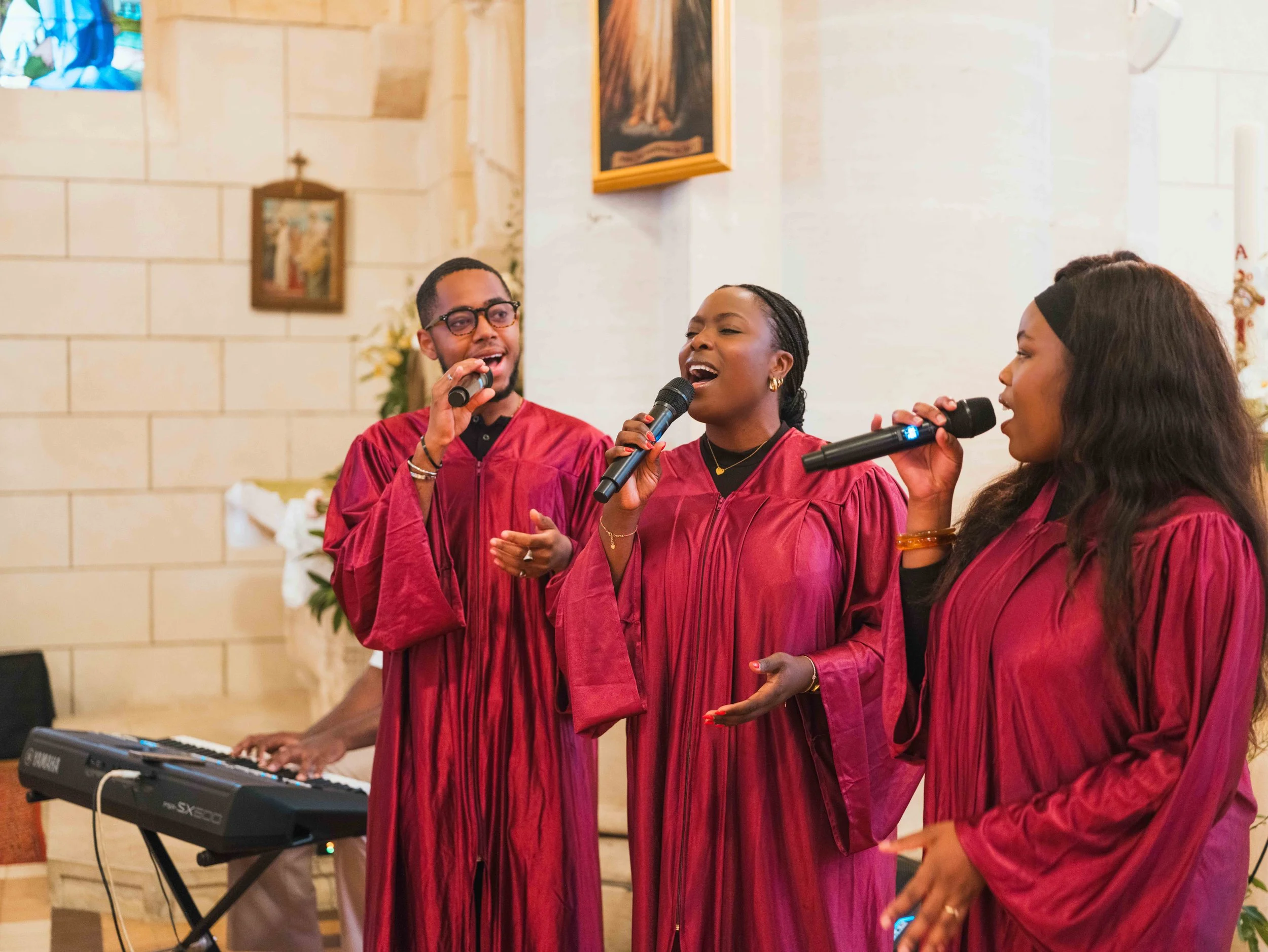 Trois choristes gospel de webelieve music chantent avec des microphones lors d'une célébration religieuse dans une église, vêtues de robes de cérémonie bordeaux.