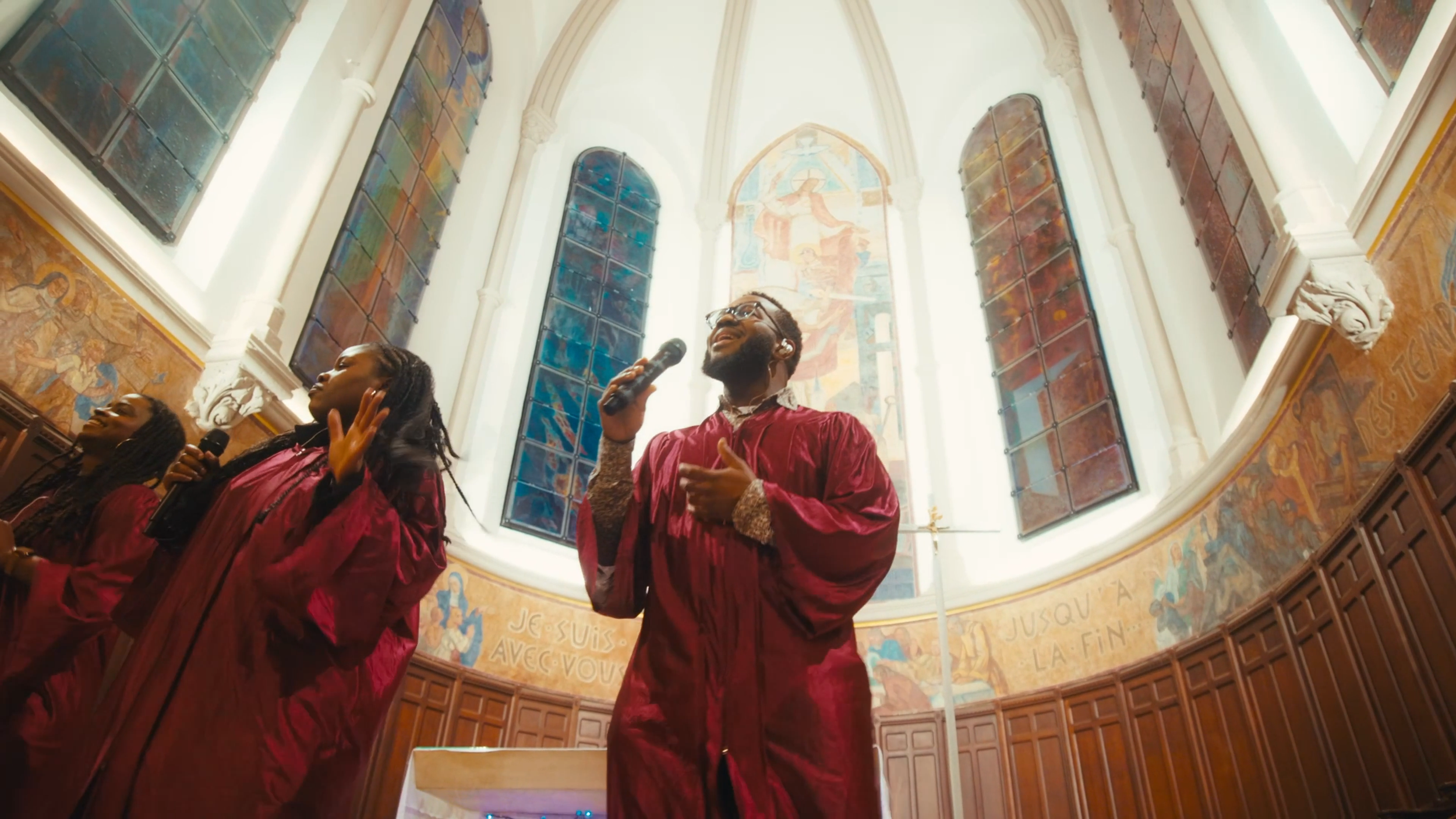 Trois choristes Webelieve gospel chantant dans une église. Le chœur porte des robes rouges, l’intérieur de l’église est décoré avec des vitraux colorés et des fresques murales.