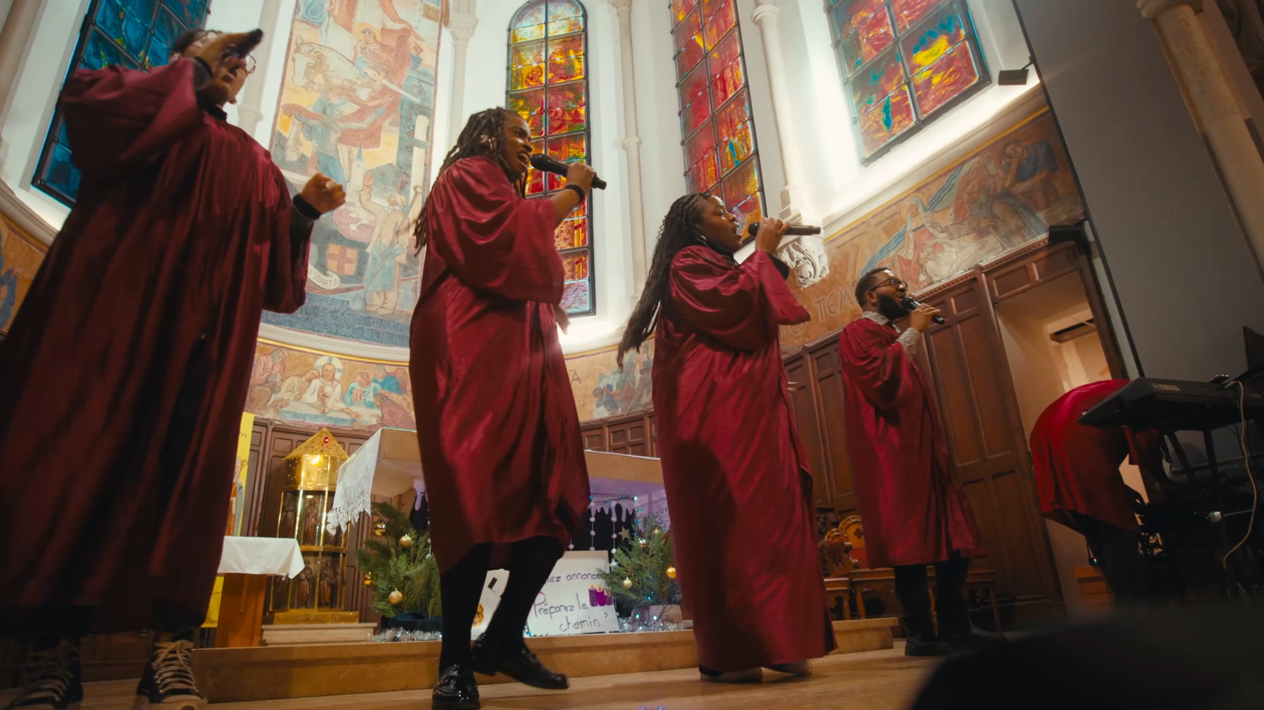 Groupe de cinq personnes chantant dans une église avec des vitraux colorés en arrière-plan, portant des robes rouges.