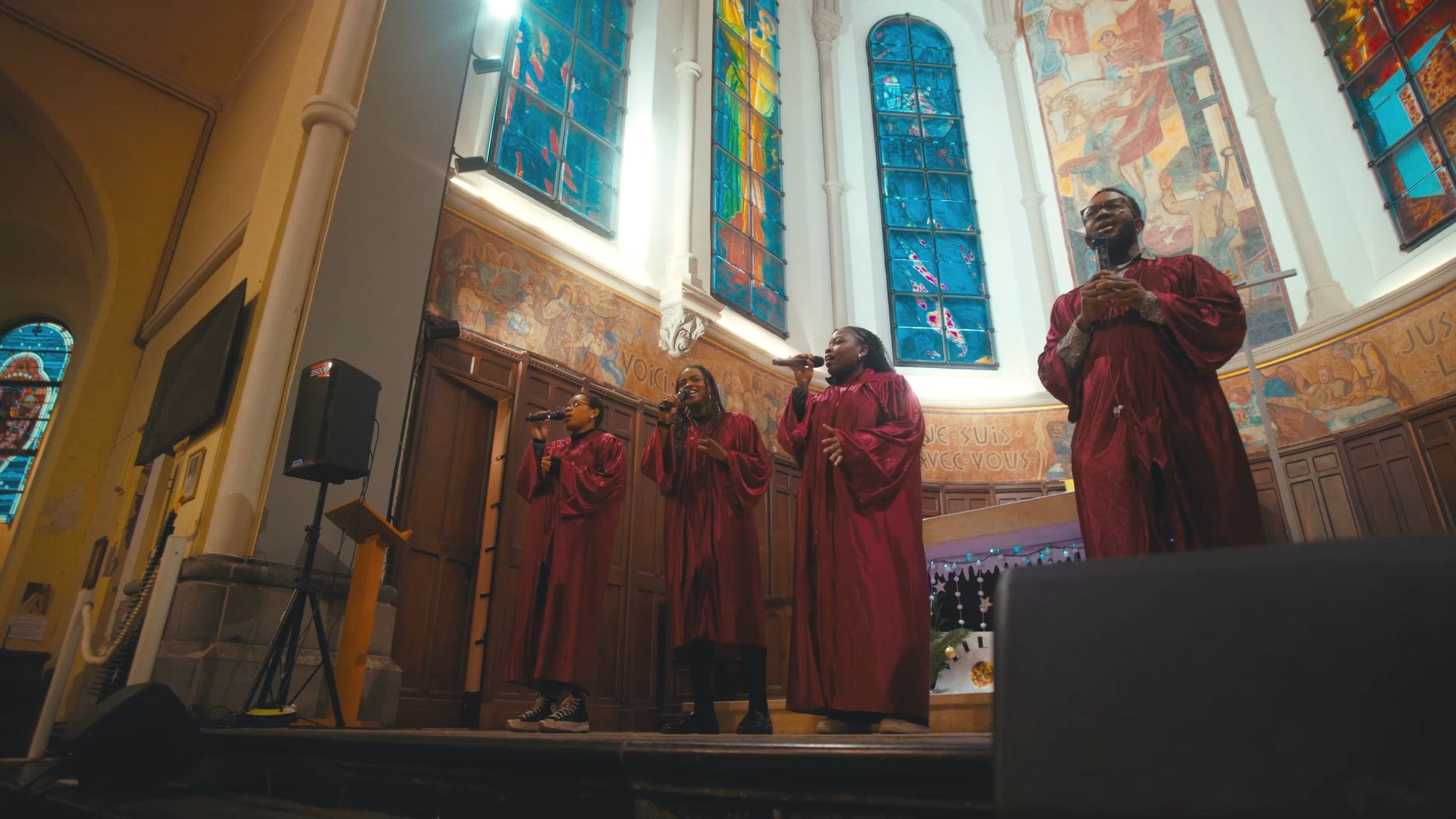 Quatre personnes chantent avec des microphones dans une église, portant des robes rouges, sous des vitraux colorés.