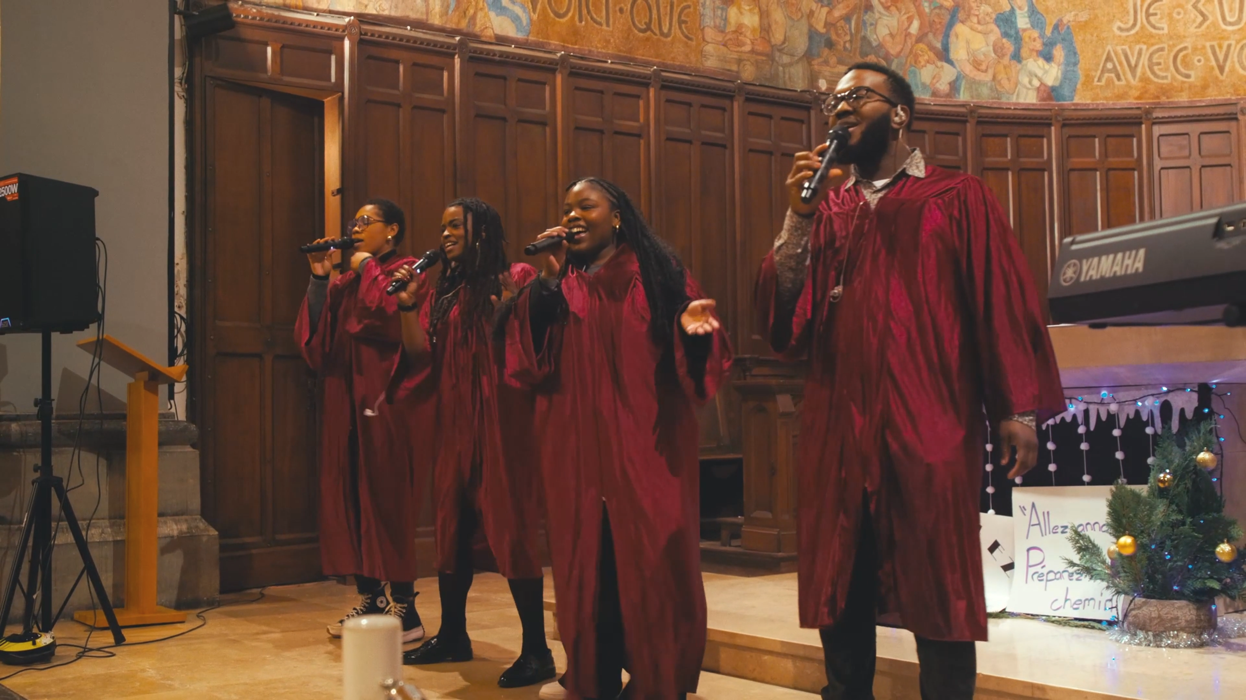 Groupe de cinq jeunes chanteurs en robe de chambre bordeaux chantant avec micro dans une église ou un lieu religieux, décoré pour Noël avec petit arbre et affiche