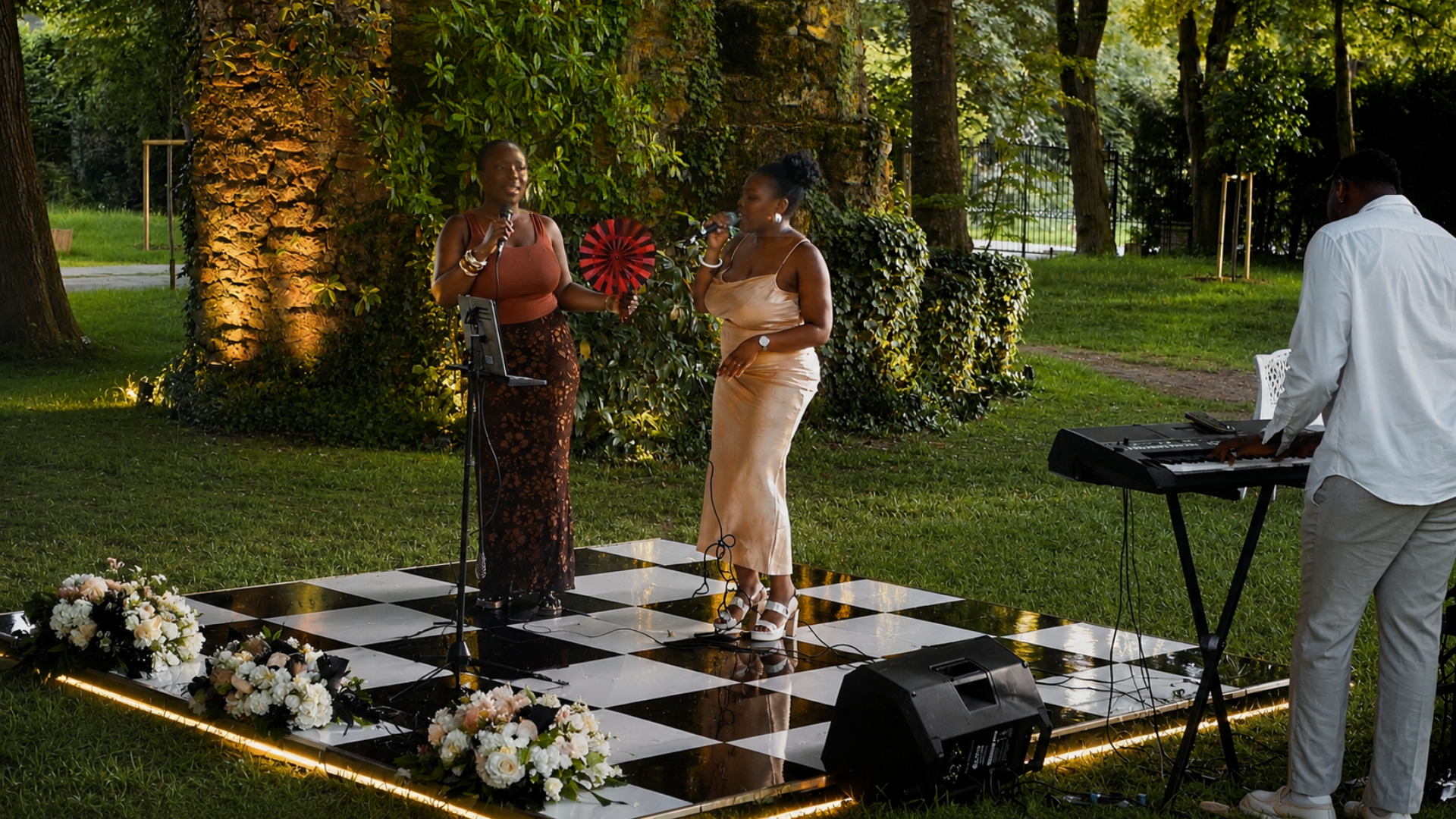 Deux femmes chantent en plein air, l'une avec un micro et l'autre avec un éventail, accompagnées par un musicien jouant du clavier sur une scène noire et blanche damier décorée de fleurs lors d'un mariage luxueux