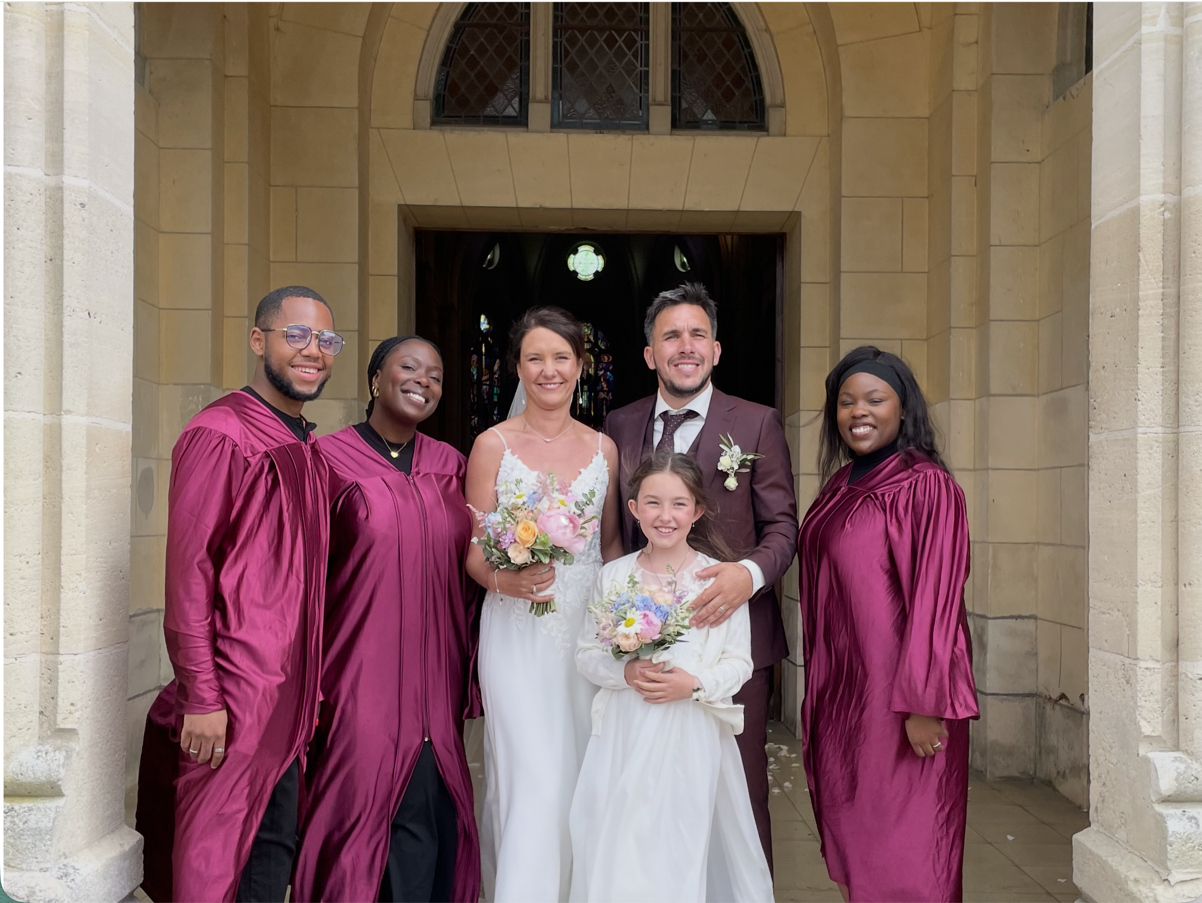 Groupe de personnes lors d'une cérémonie de mariage devant une église en pierre avec des vitraux colorés, portant des robes de cérémonie et des costumes, souriant pour la photo.