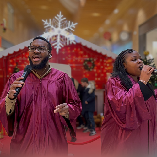 Deux personnes chantent et tiennent des microphones, portant des robes de graduation, avec décoration festive de Noël en arrière-plan.