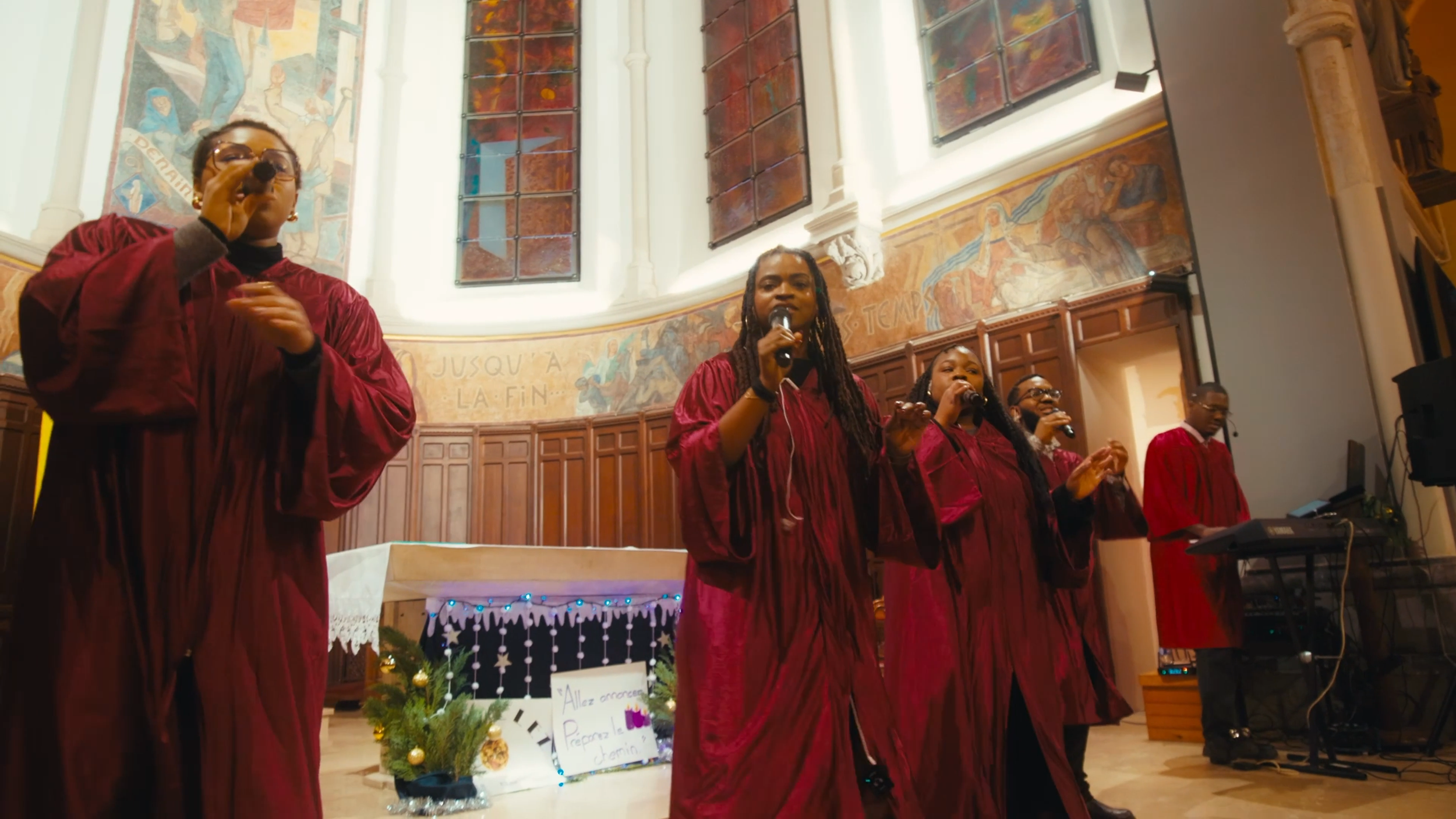 Groupe de personnes en robe de graduation rouge chantant dans une église, avec des vitraux et une décoration festive en arrière-plan.