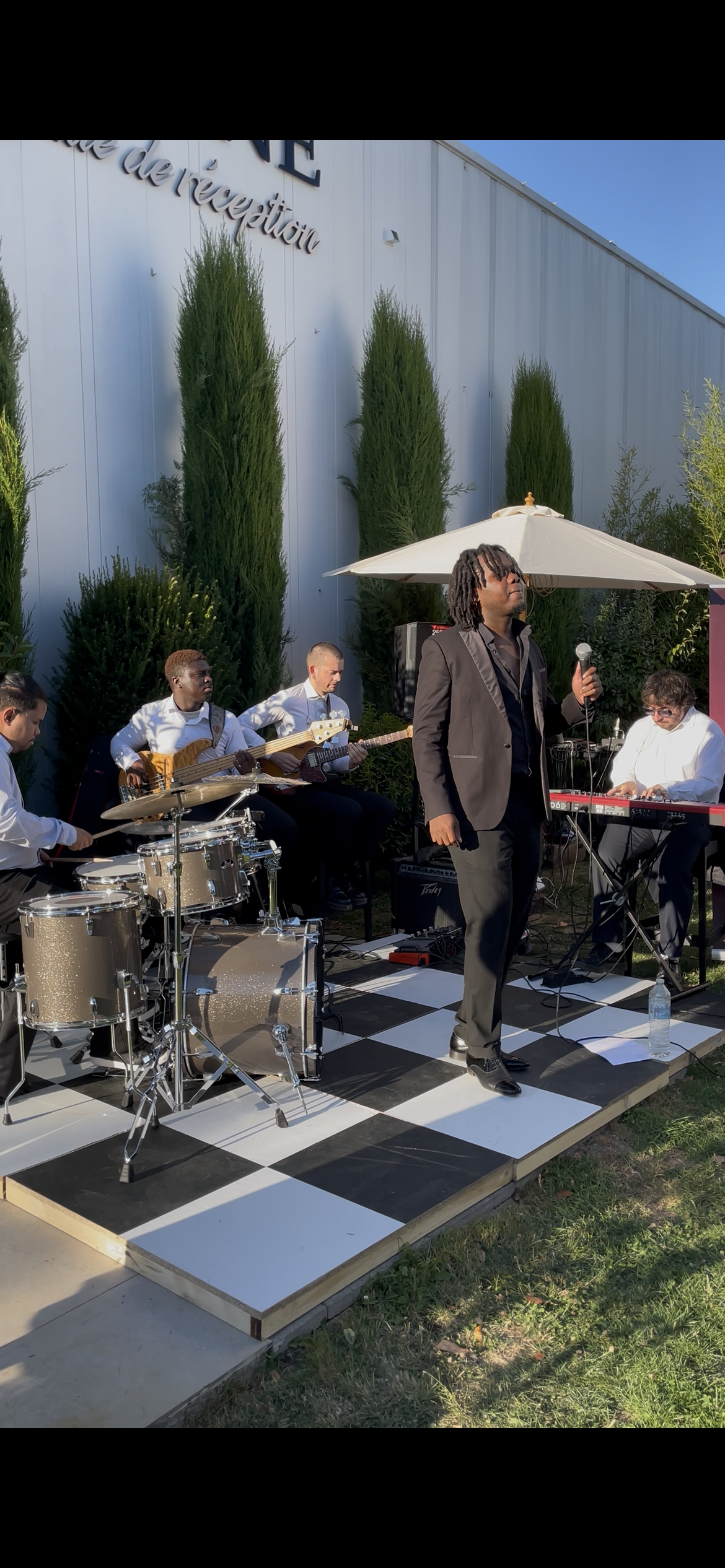 Un groupe de musiciens Webelieve jouant lors d'une performance en plein air. Il y a un chanteur avec un micro, un clavier, un guitariste, un bassiste et un batteur. La scène est sous un grand parasol,