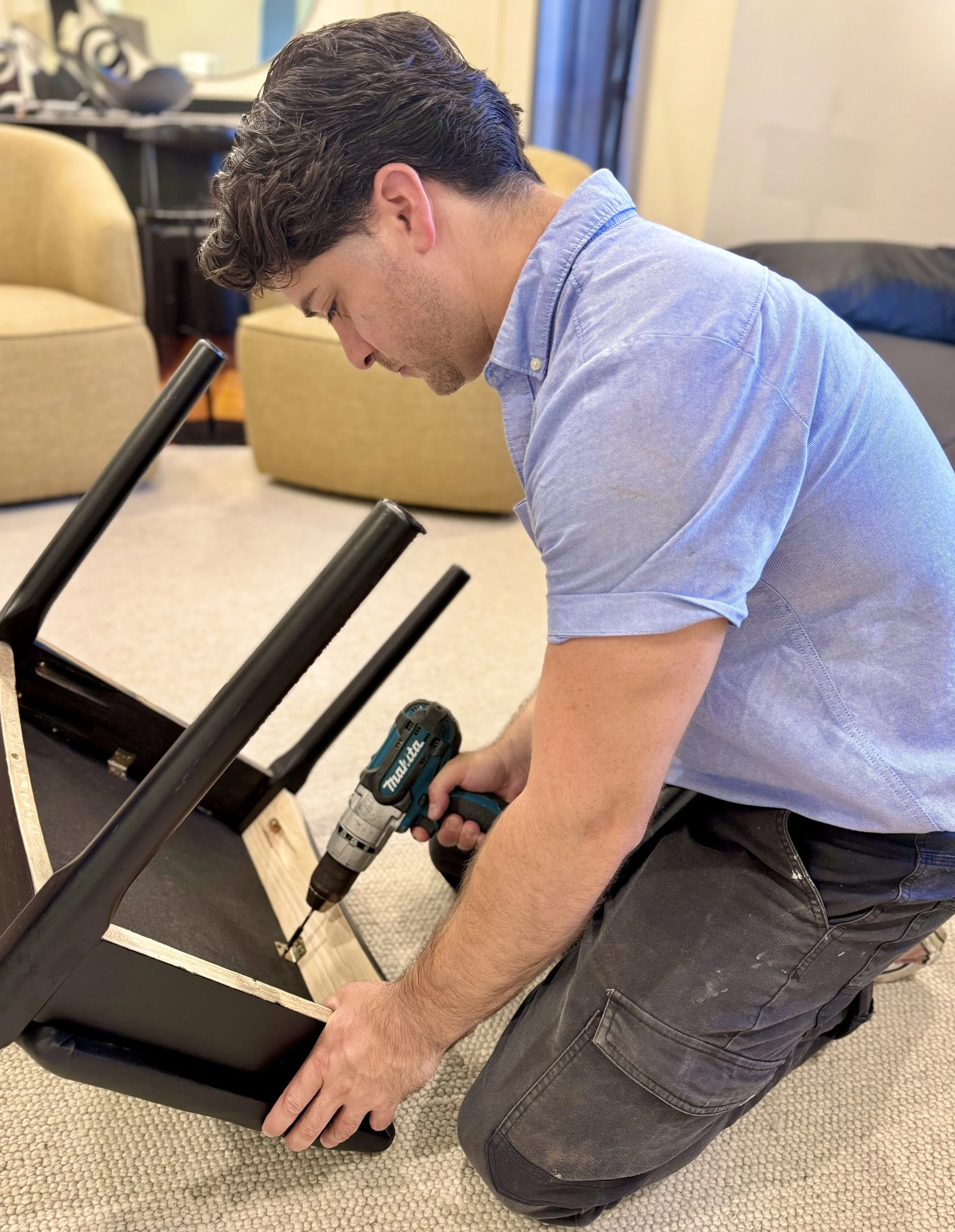 Jake's Maintenance assembling a black chair with a cordless drill in a living room.