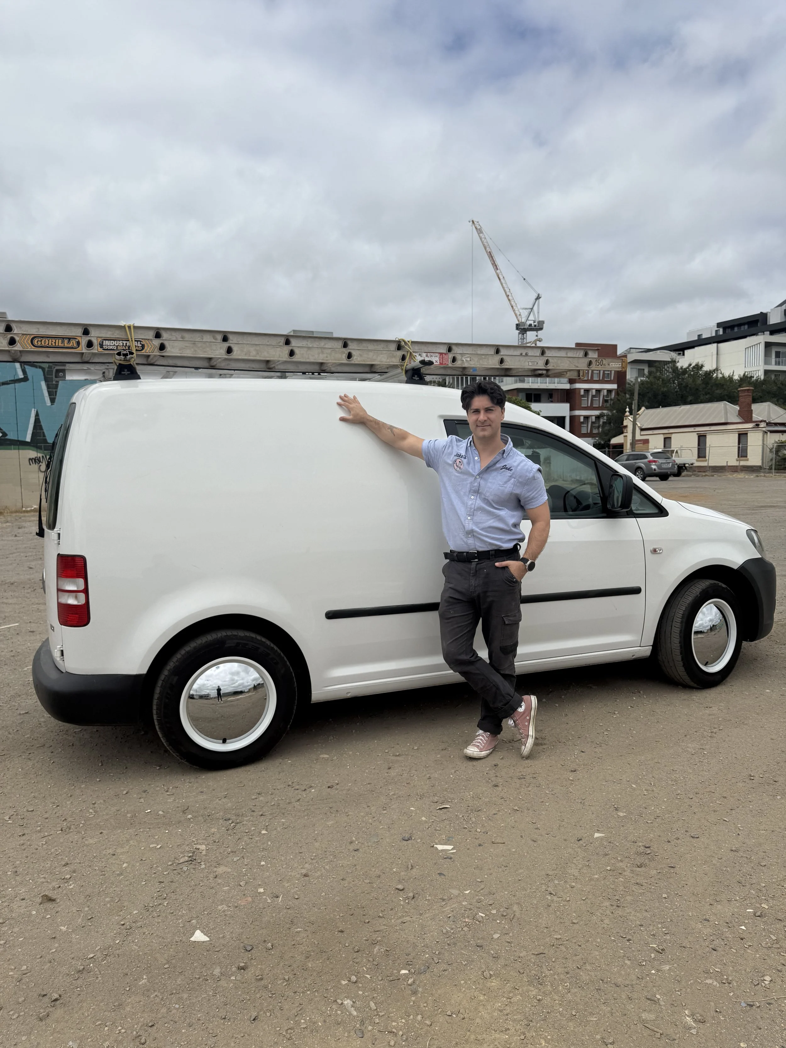 Jake's Maintenance wearing a light blue shirt and black pants standing next to a white cargo van with a ladder on top, in an outdoor lot with buildings and a crane in the background.