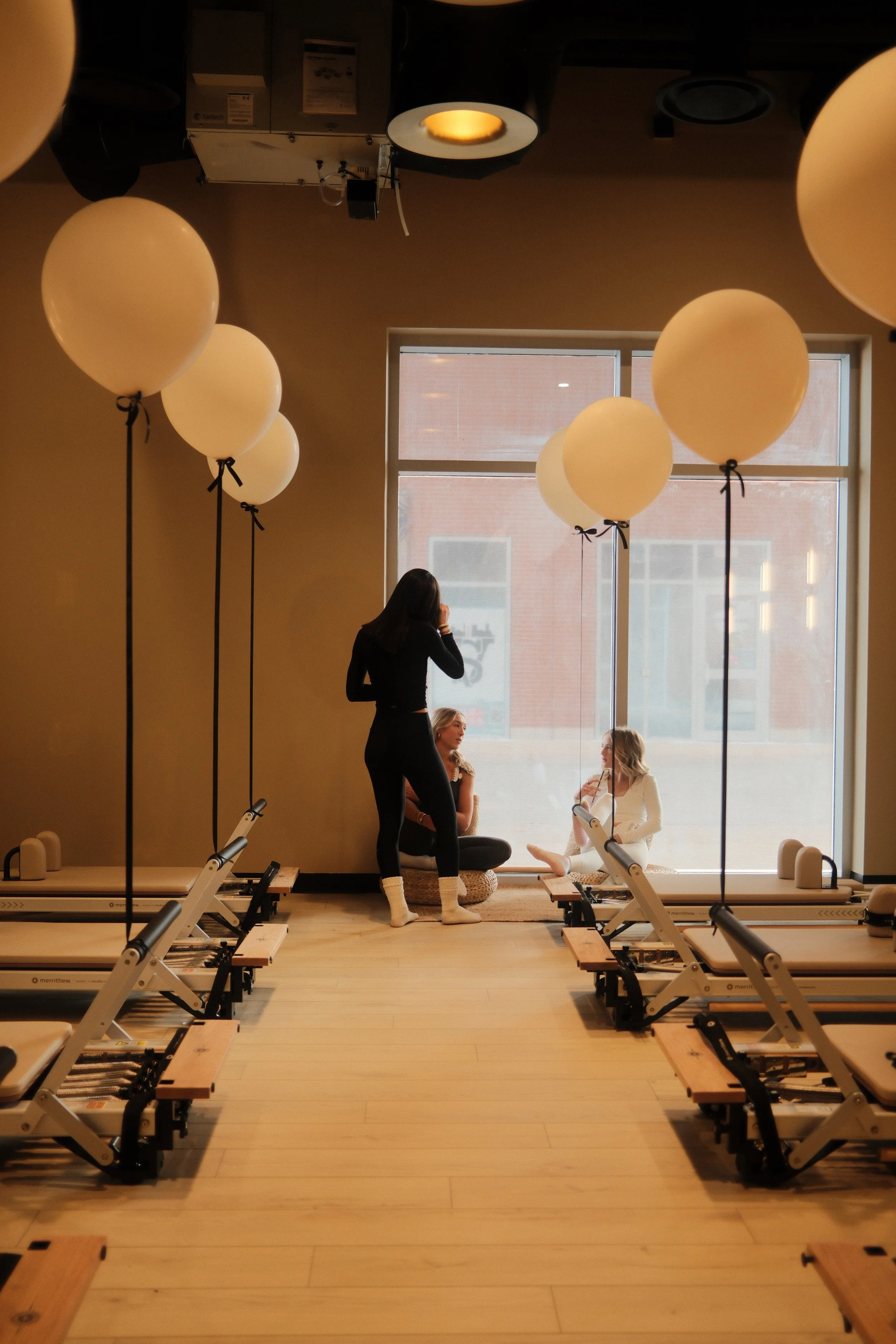 Three women in a Pilates studio, with two sitting on the floor near the window and one standing with her back to the camera, surrounded by white balloons tied to supports, and Pilates reformer machines lined up on each side of the room.