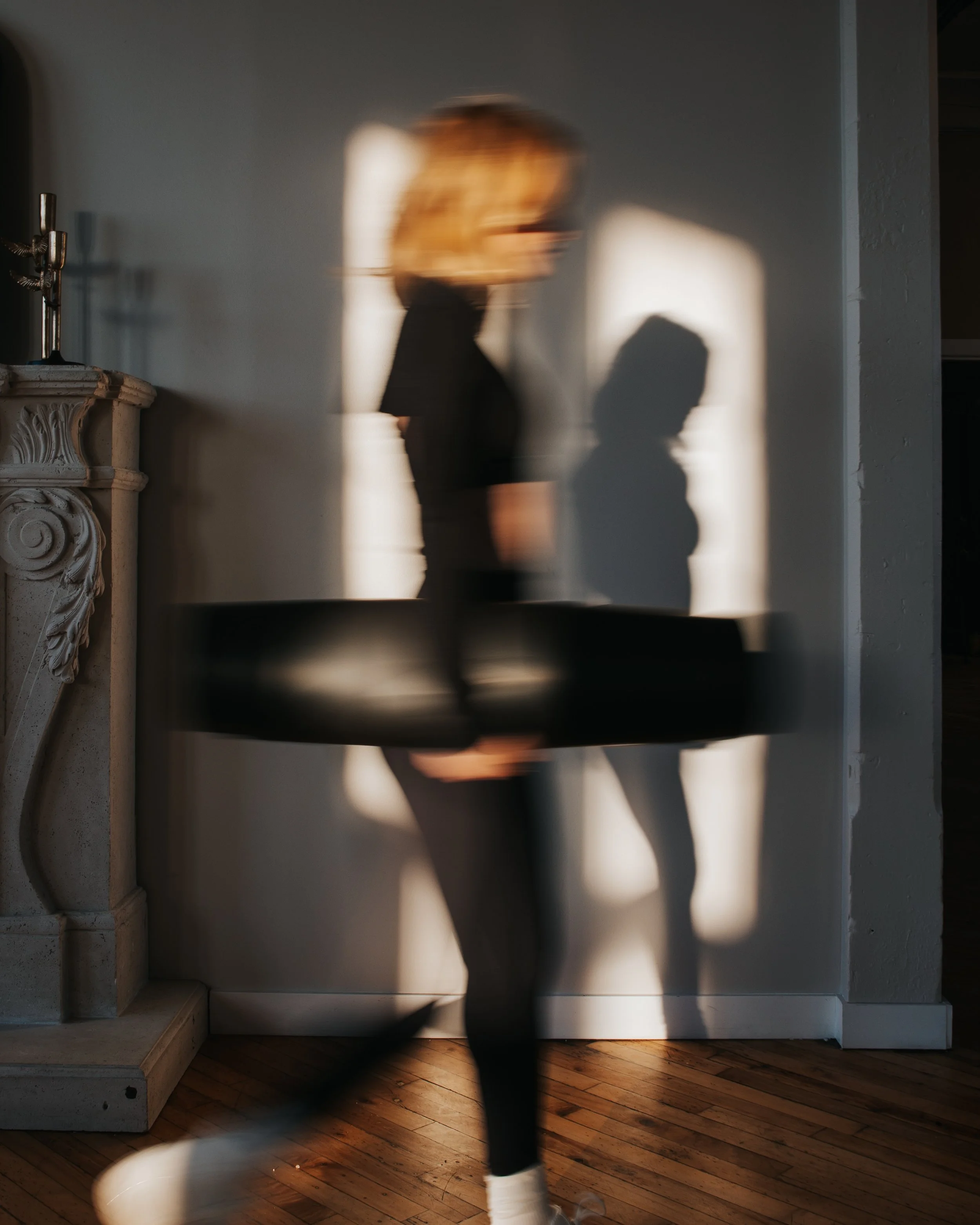 A woman holding a black platter in motion, walking past a wall with sunlight casting shadows, including her own silhouette, in a room with wooden floors and a decorative fireplace.