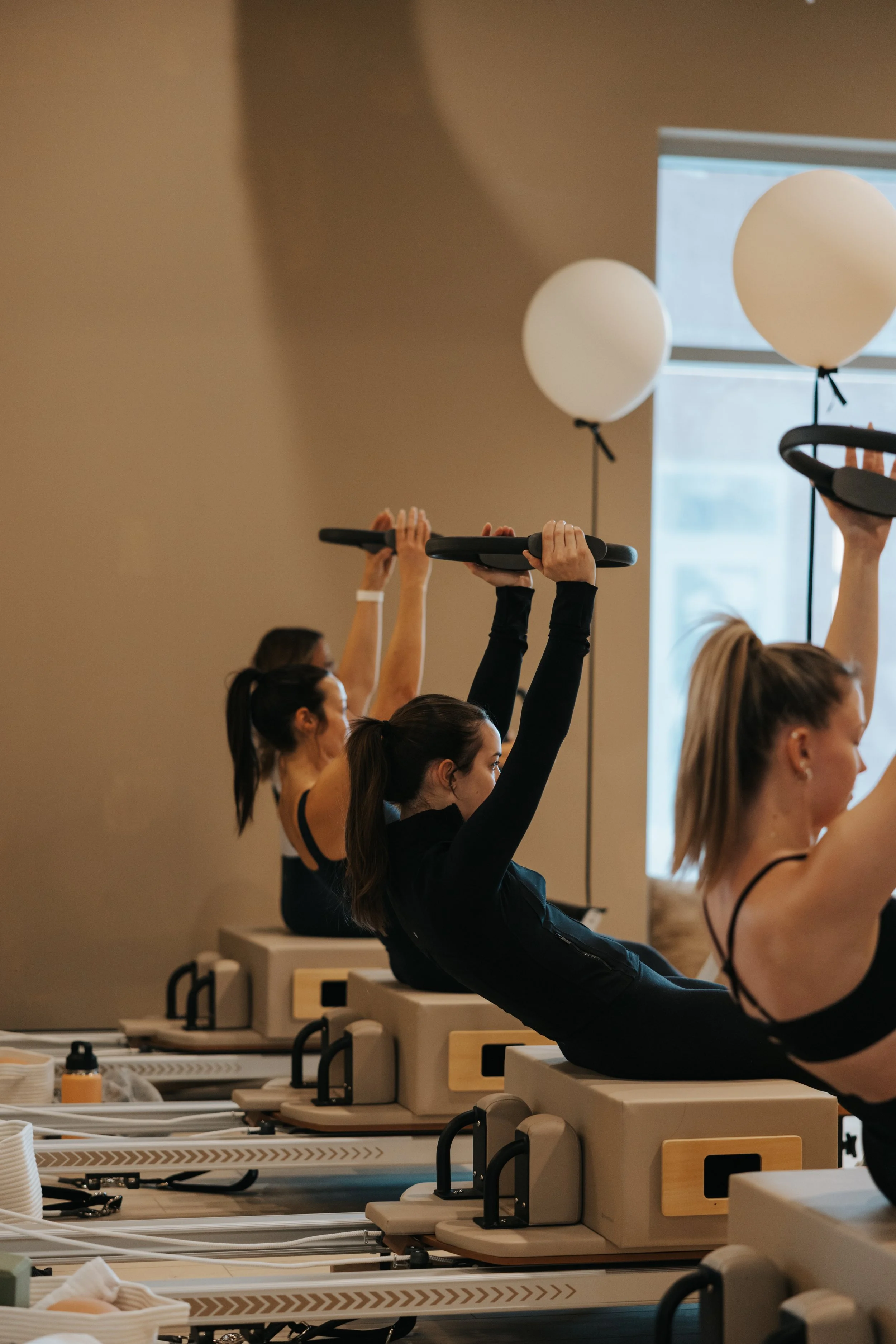 Women participating in a fitness class, lifting black exercise bars with white balloons in the background.