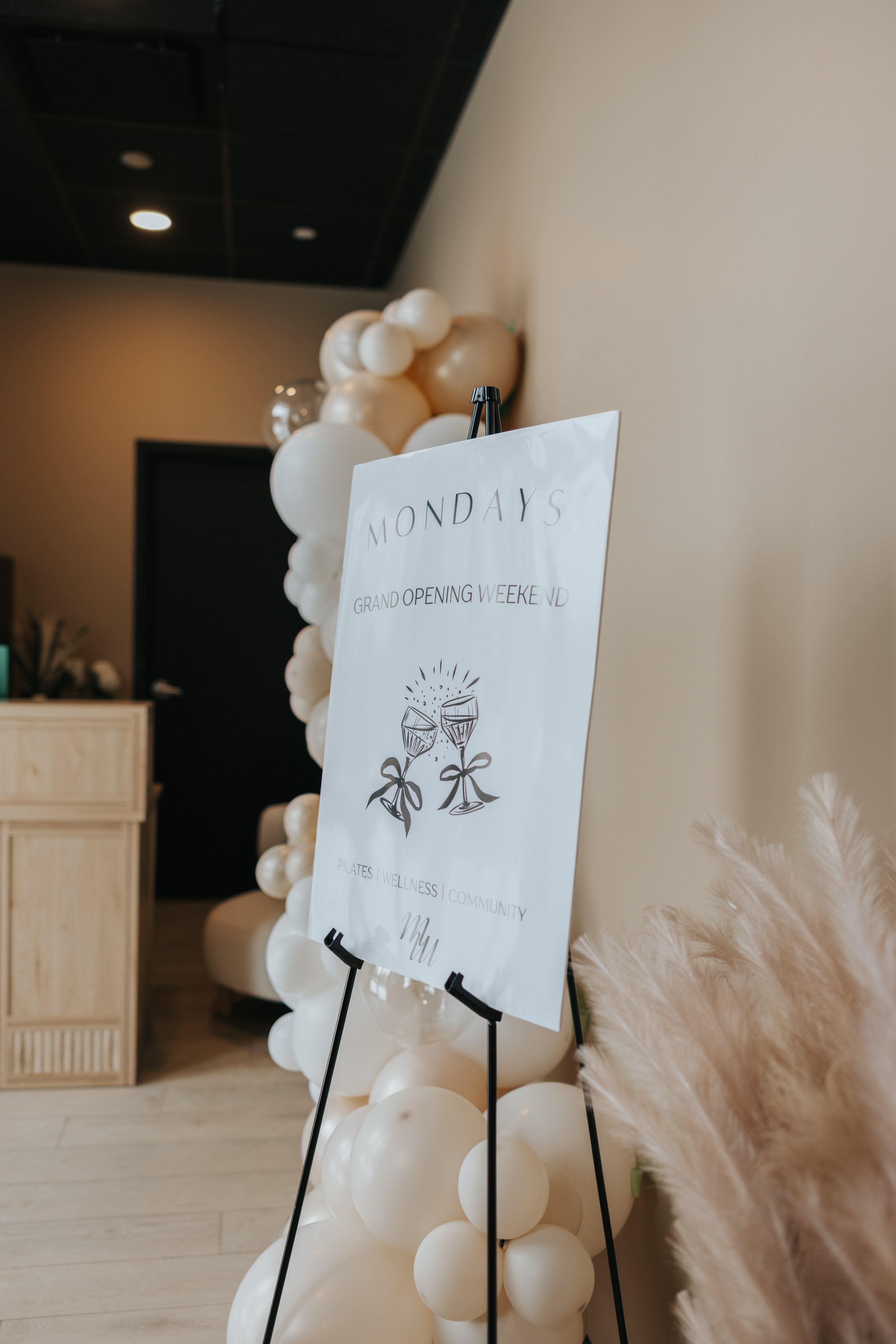 Event sign on easel with balloon arch in background, promoting grand opening weekend with activities like Pilates, wellness, and community.