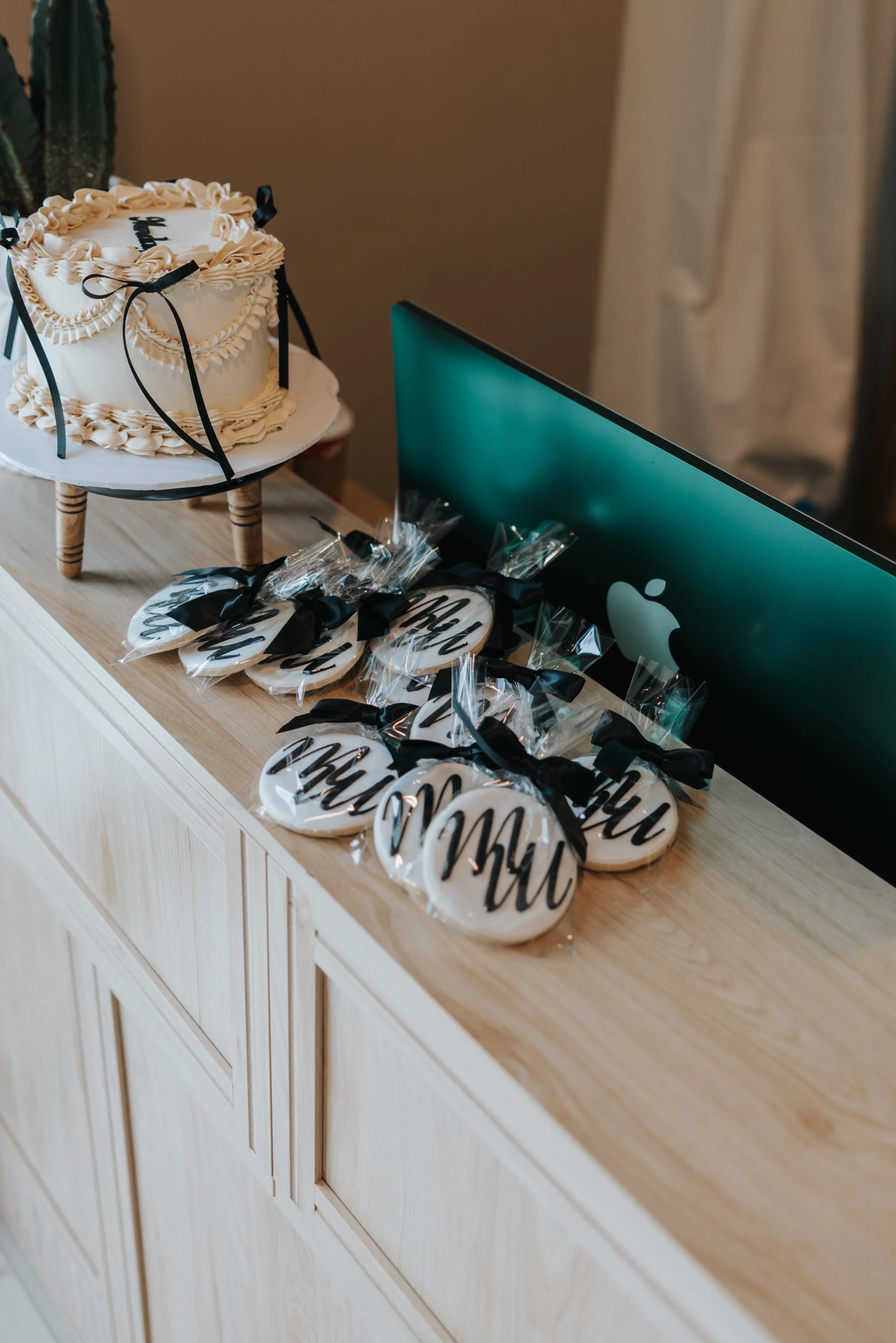 A cake decorated with white frosting and black ribbons, placed on a small wooden stand, is on a wooden sideboard. Next to it, there are several round cookies wrapped in clear plastic with black ribbons, featuring the word "Mum" in black script.