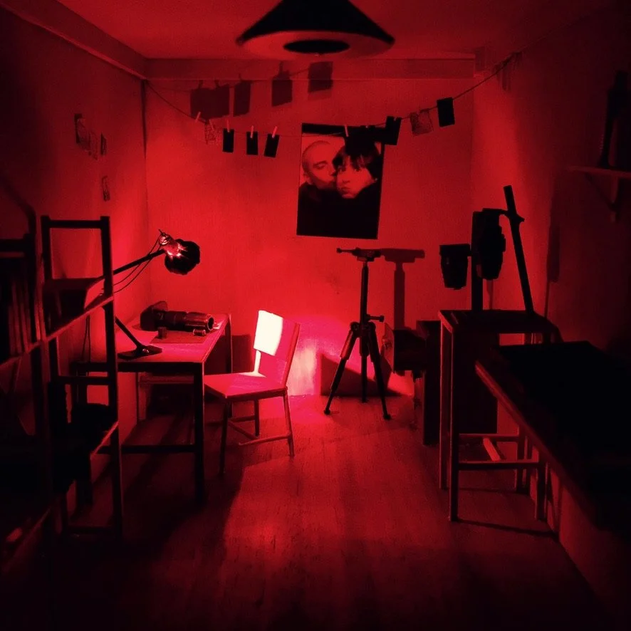 Een kamertje met een roodgekleurde verlichting, een tafel met een stoel, een verstelbare bureaulamp, een camera op statief, een verrekijker, een poster op de muur en wat hangende foto's of briefjes.