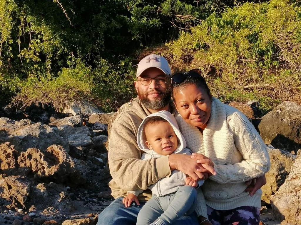 A family of three sitting outdoors on rocks near greenery, with the sun shining, including a man, a woman, and a young child.