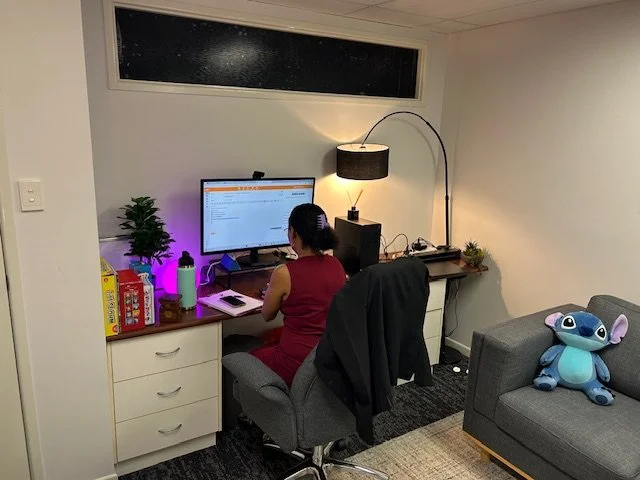 Woman sitting at a desk working on a computer with a monitor, books, plants, and a purple light in a living room with a gray couch and a stuffed Stitch toy