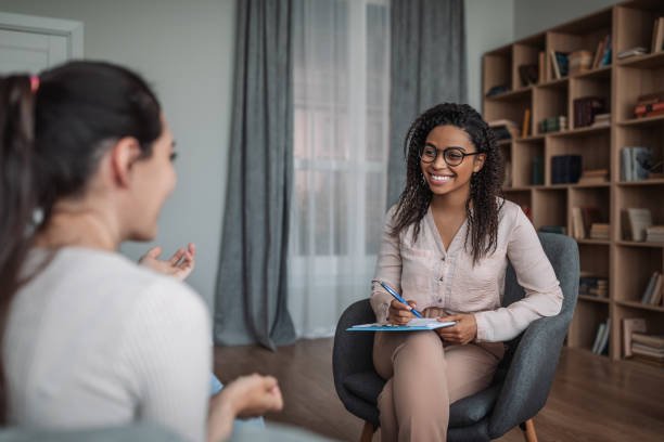 Two women having a conversation in an office setting, one with curly hair and glasses, holding a notepad and pen, smiling at the other woman.