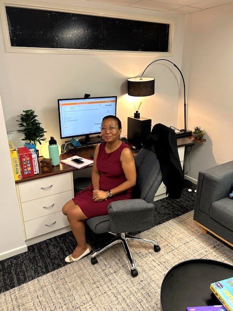 Woman sitting at a desk with a computer monitor, desk lamp, and various office supplies in a small office or study.
