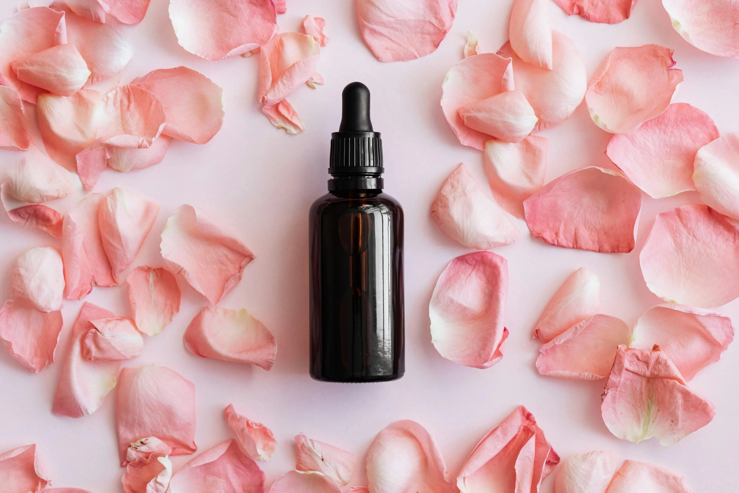 A dark glass dropper bottle surrounded by pink rose petals on a light pink background.