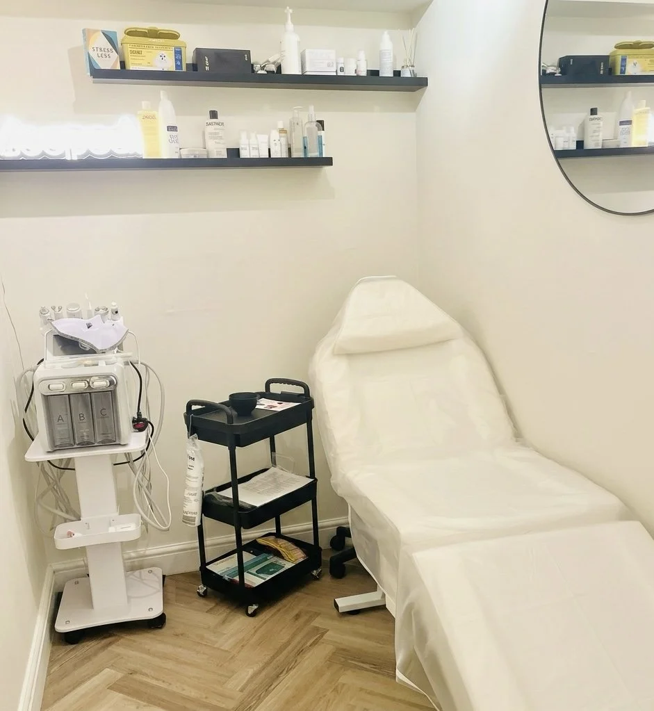 Medical examination room with an adjustable examination table covered with white paper, medical supplies on shelves, a small rolling cart, and a piece of medical equipment with tubes.