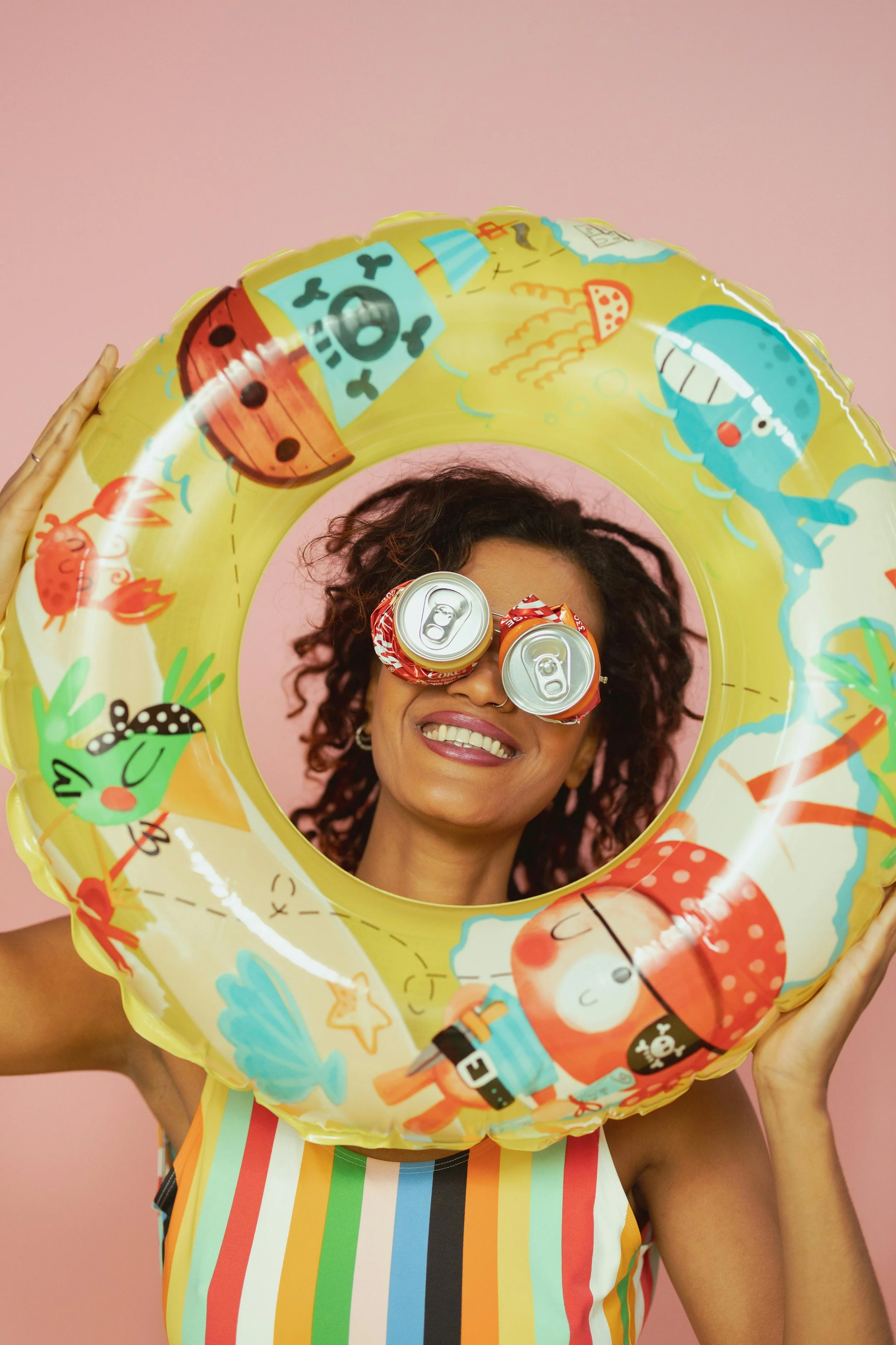 A woman with curly hair and a big smile holding a colorful inflatable ring with pirate and ocean creature illustrations in front of her face, with soda cans as glasses, against a pink background.