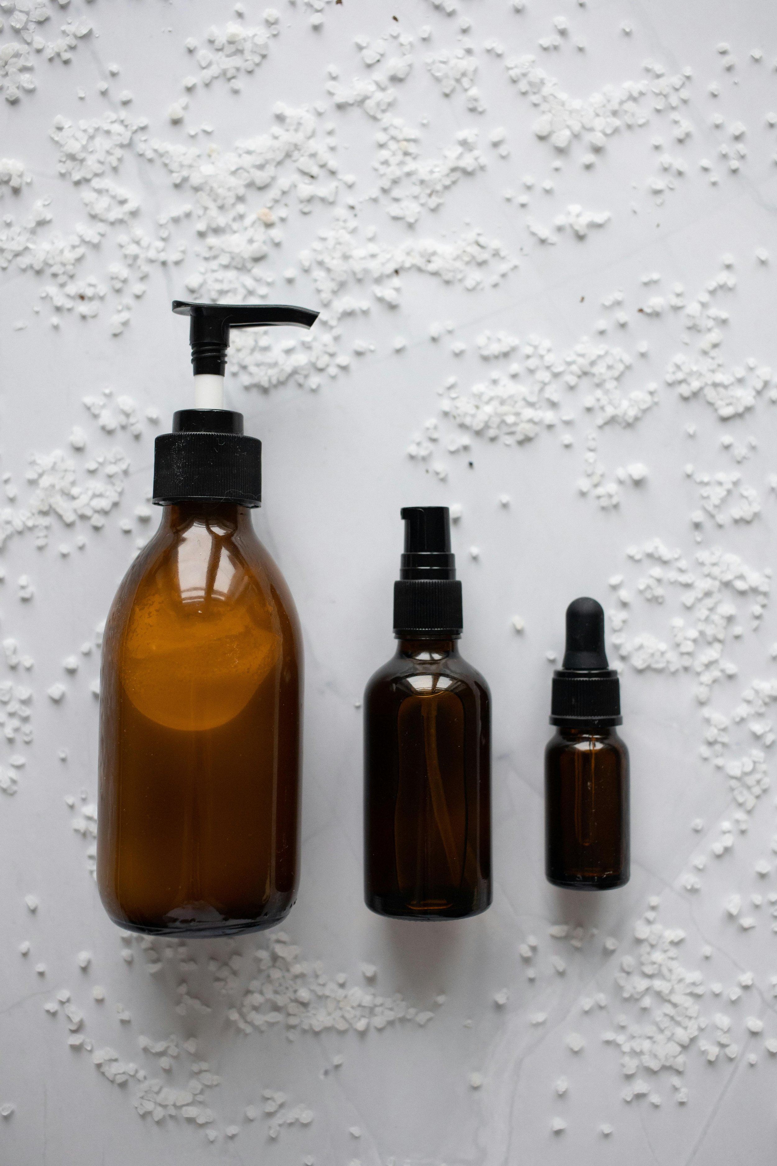 Three amber glass bottles with black pump dispensers are surrounded by scattered white grains or small particles on a white surface.