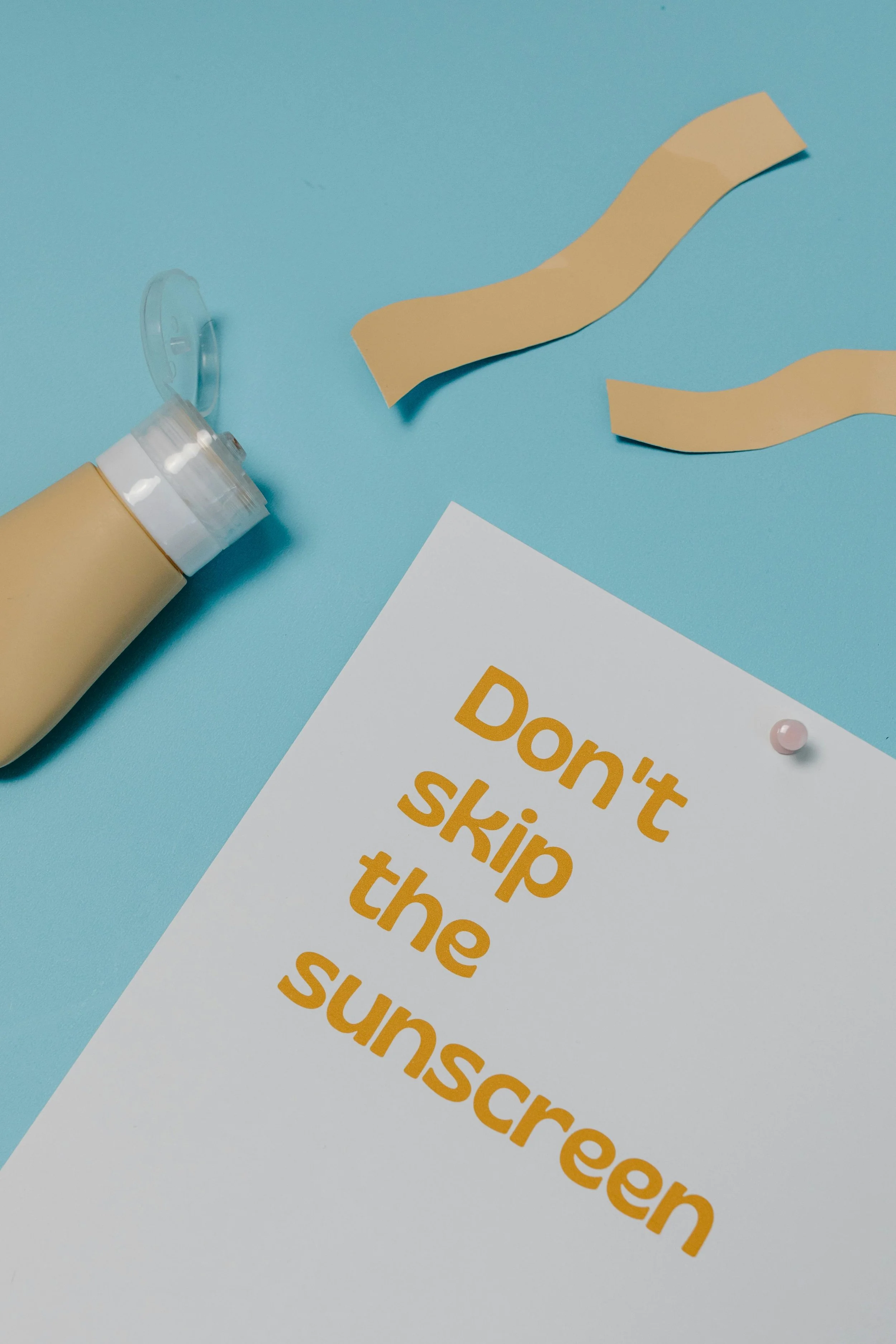 A white sheet of paper with the yellow print text 'Don't skip the suncreen' on a light blue background. A yellow sunscreen bottle with the cap open, two beige paper strips, and a pink push pin are also on the background.