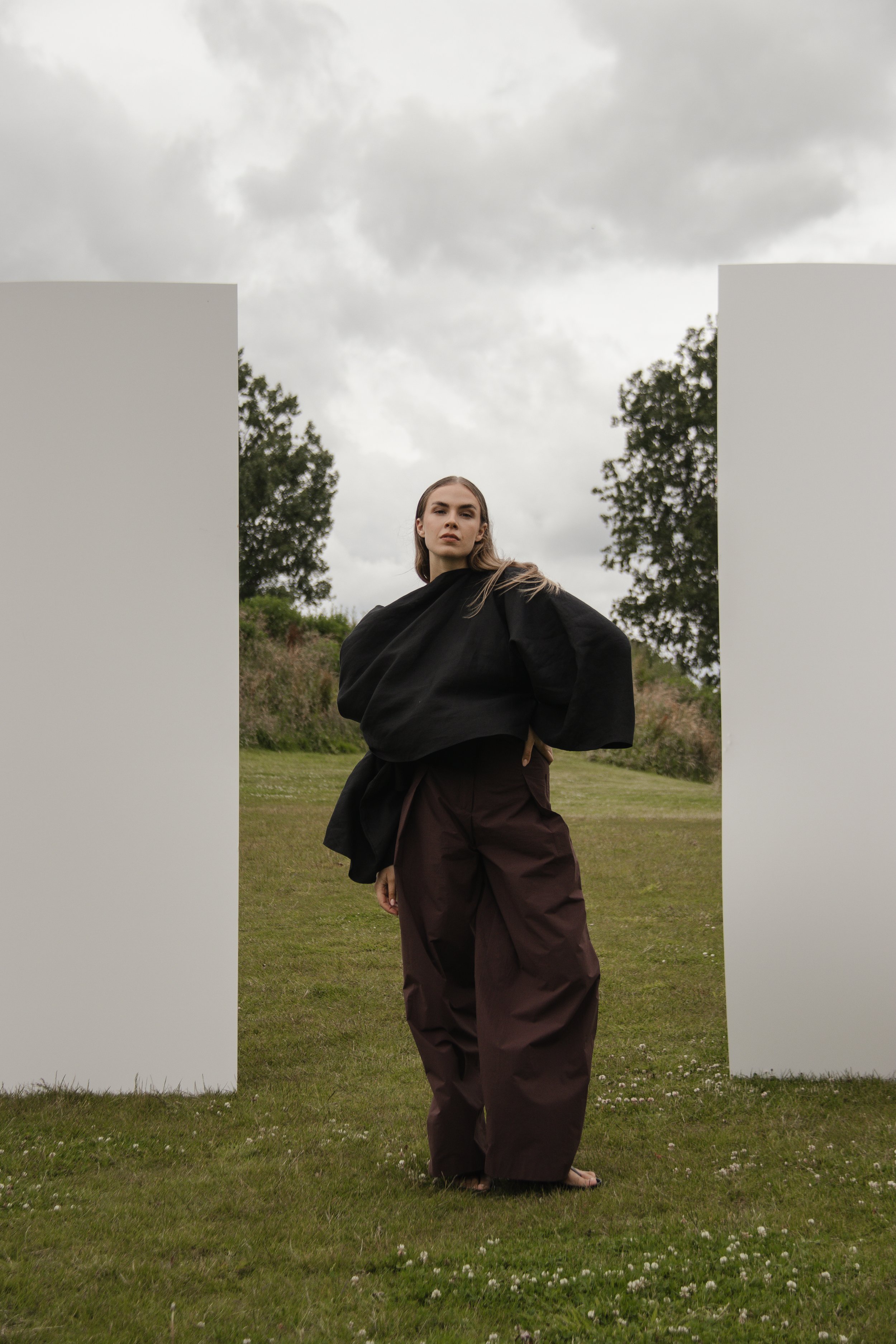 A woman standing outdoors on a grassy field between two white panels, wearing a black oversized top and brown wide-leg pants, under a cloudy sky.