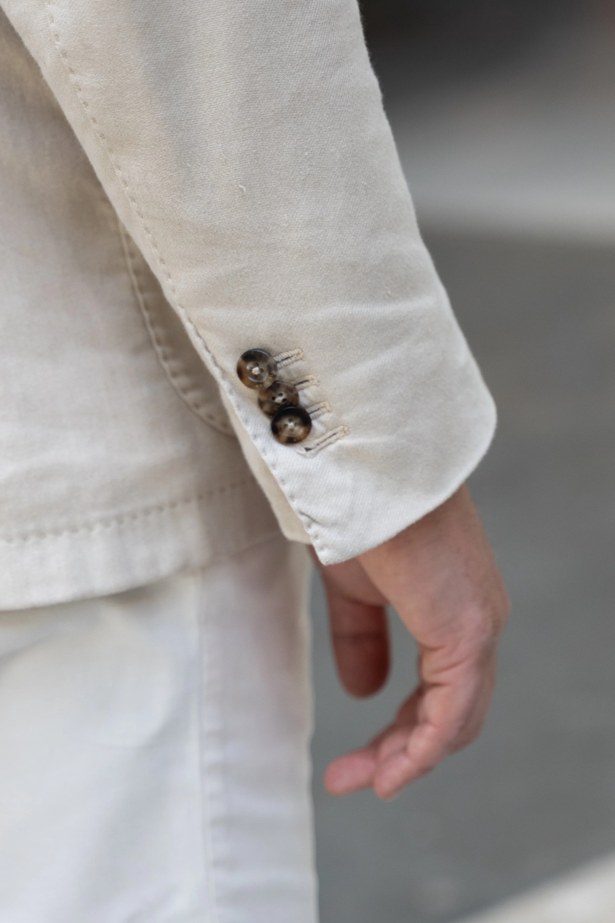 Close-up of a light-colored jacket sleeve with three dark buttons near the cuff, worn by a person with a beige shirt and white pants.